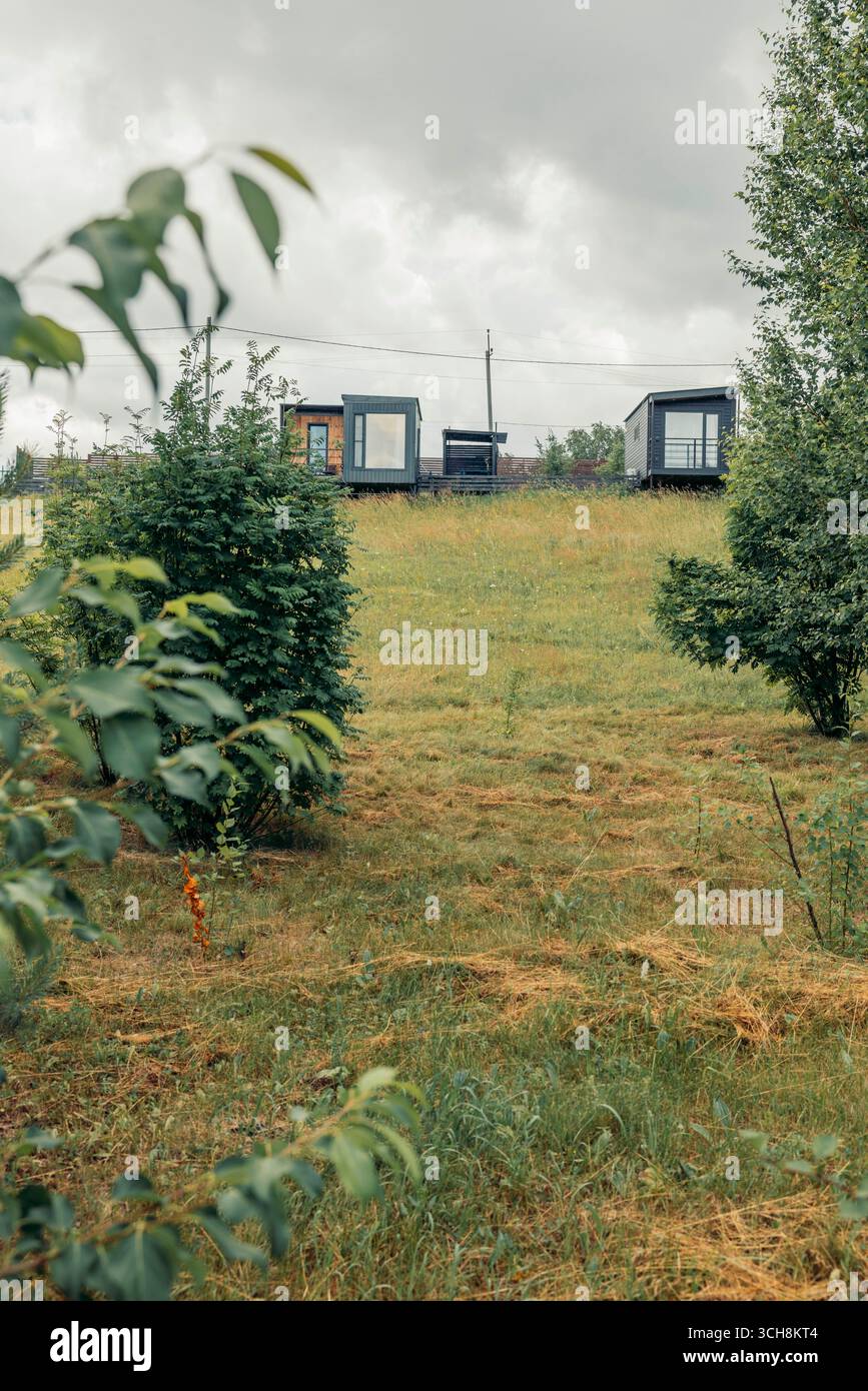 Moderne modulare Häuser mit großen Fenstern, auf grünen Hügeln, Gras und Büsche unter bewölktem Himmel, zeitgenössische Architektur für Öko-Wohnen, Landschaft Stockfoto