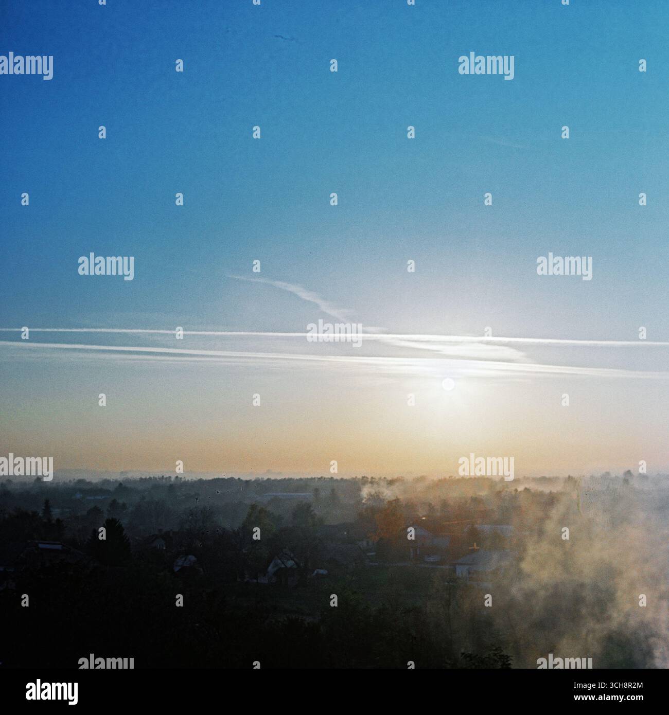 Luftaufnahme eines Dorfes der Vojvodina an einem sonnigen Wintermorgen, mit heißem Rauch und Sonne, die in die Linse am klaren blauen Himmel, aufgenommen auf Farbfilm. Stockfoto