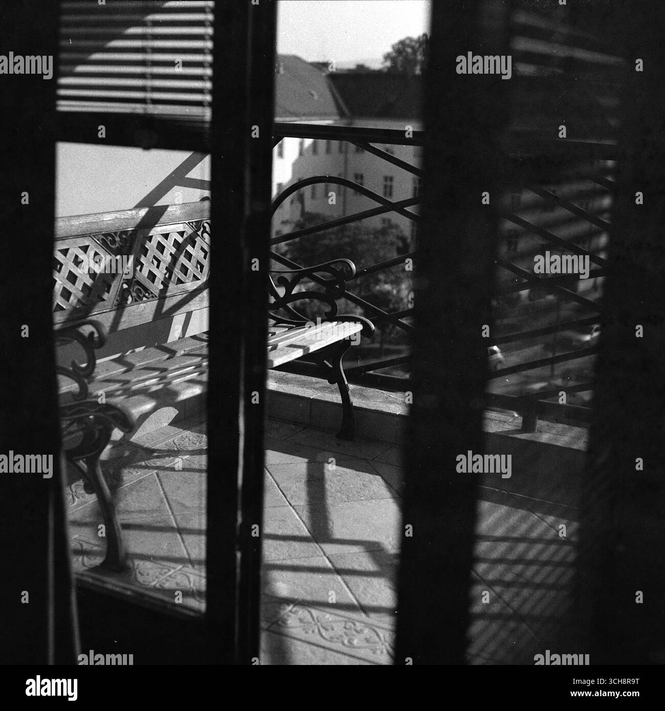 Durch eine Terrassentür geschossen: Eine leere Bank auf einer sonnigen Terrasse mit Geländerschatten, in Schwarz-weiß auf einer Hasselblad festgehalten. Stockfoto