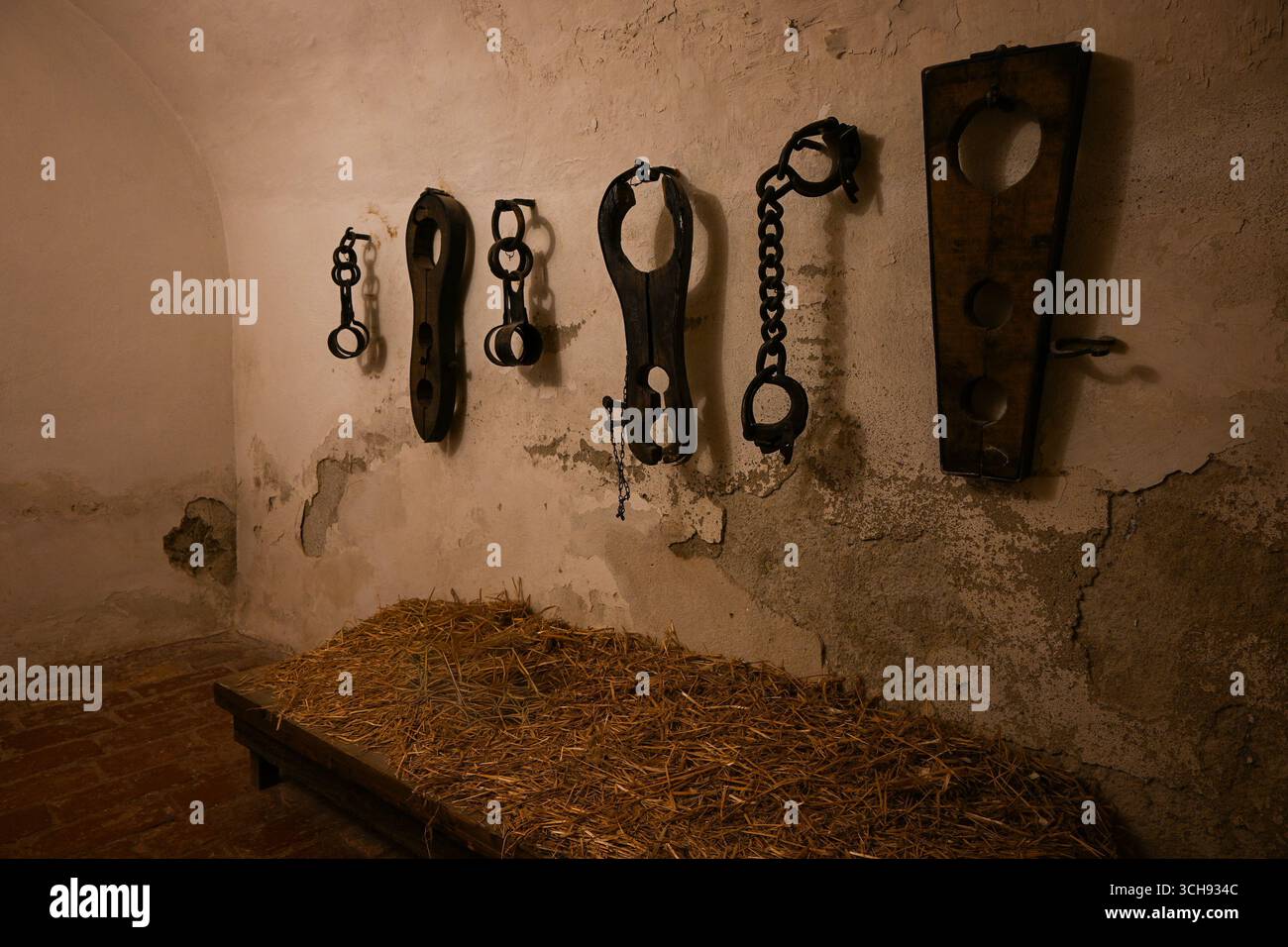 Verschiedene Foltergeräte hängen an einer verfallenen Mauer in einem mittelalterlichen Kerker mit Stroh auf einem Holzbett. Stockfoto