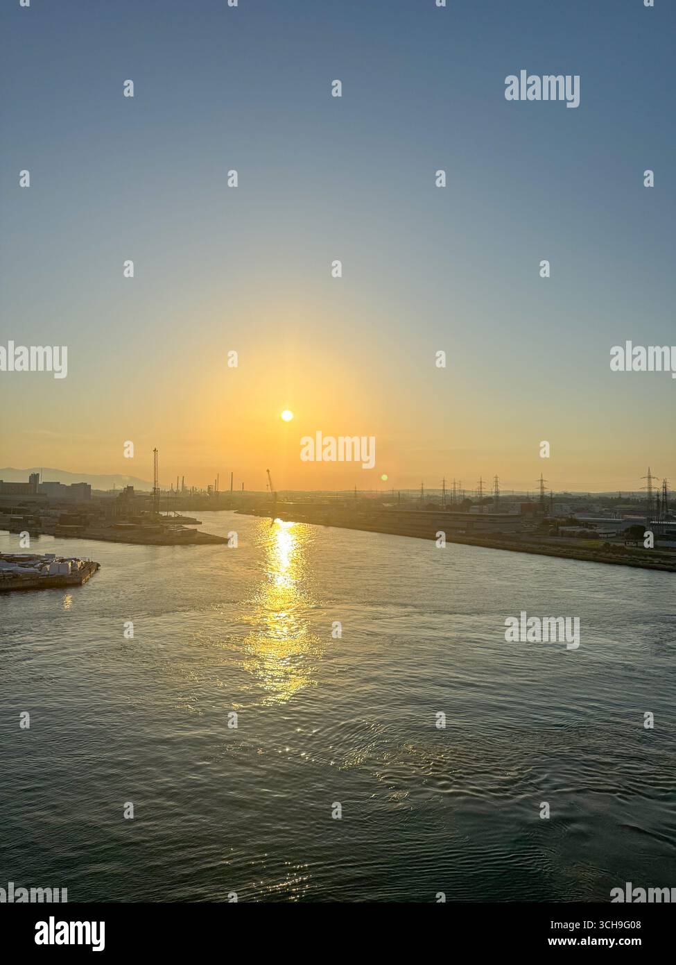 Ruhiger Sonnenuntergang über einem ruhigen Fluss mit goldener Reflexion auf der Wasseroberfläche. Im Hintergrund sind die Industriestrukturen dargestellt Stockfoto