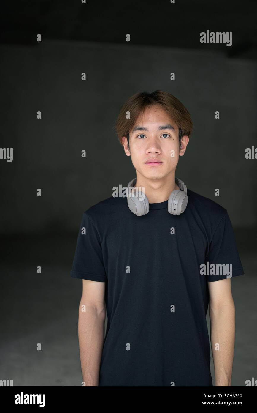 Junger thailändischer asiatischer Mann mit einem schwarzen T-Shirt, posiert in einem minimalistischen, brutalistischen Baubereich mit grauen Betonwänden Stockfoto