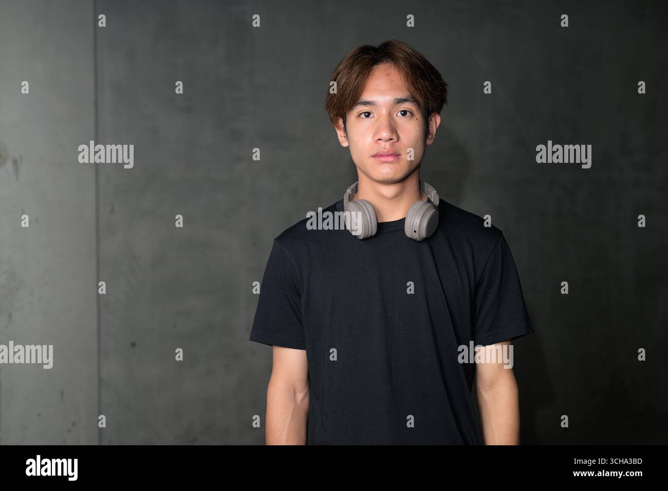 Junger thailändischer asiatischer Mann mit einem schwarzen T-Shirt, posiert in einem minimalistischen, brutalistischen Baubereich mit grauen Betonwänden Stockfoto