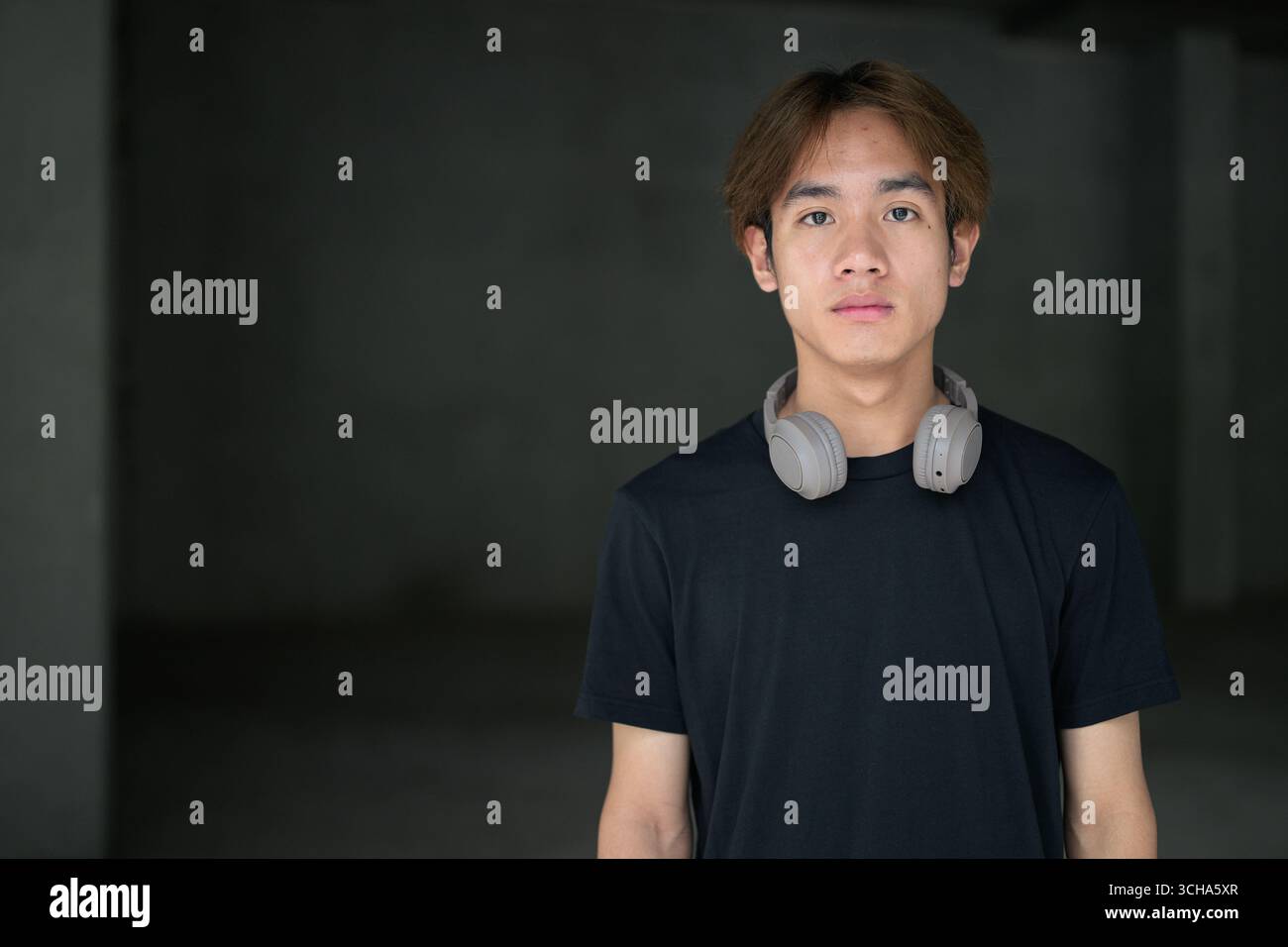 Junger thailändischer asiatischer Mann mit einem schwarzen T-Shirt, posiert in einem minimalistischen, brutalistischen Baubereich mit grauen Betonwänden Stockfoto