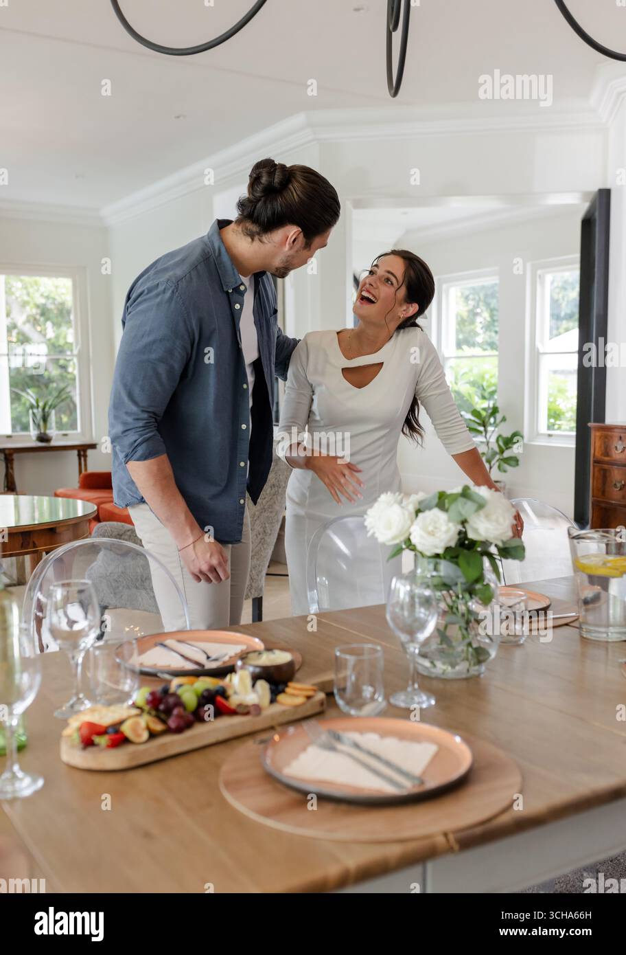 Ein Paar lacht zusammen am Esstisch mit eleganter Tischdekoration und Blumen, zu Hause. Romantik, Feier, Glück, Lachen, Intimität, Tablewa Stockfoto