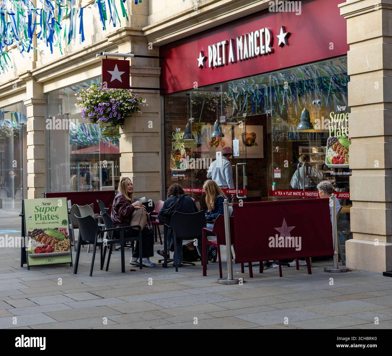Außenterrasse von Pret a Manger mit Leuten, die an einem sonnigen Tag Speisen und Getränke genießen, neben einer Ladenfront mit hängenden Blumen Bath UK Stockfoto