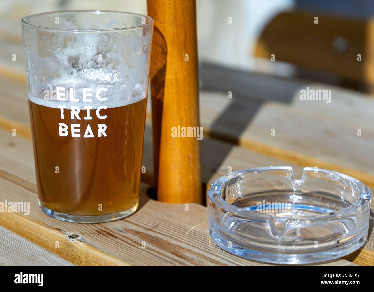 Ein halbes Glas Bier auf einem hölzernen Picknicktisch neben einem leeren Glasaschenbecher im Sonnenlicht Stockfoto