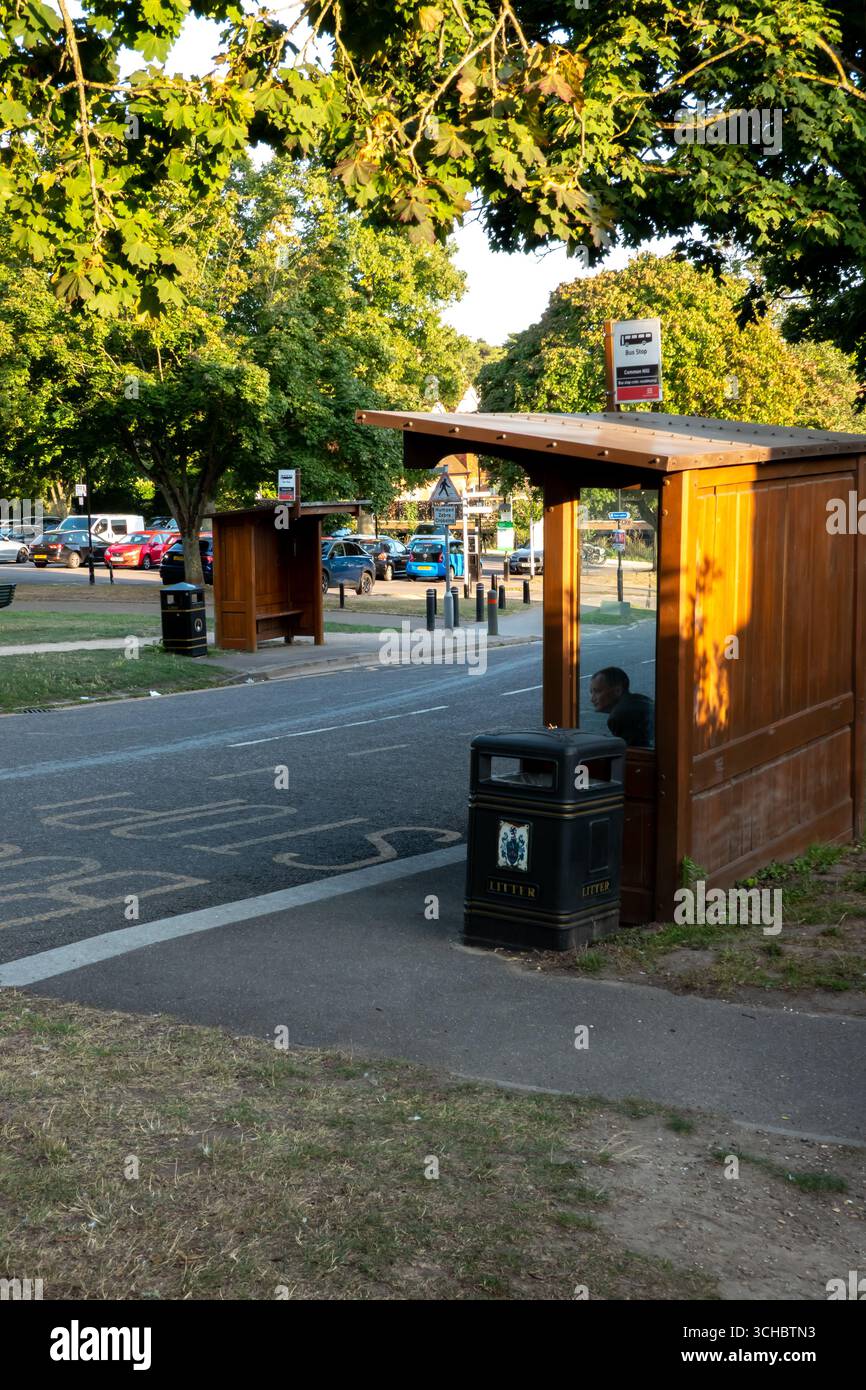 Eine Person, die in einer hölzernen Busunterkunft an einer Bushaltestelle in Saffron Walden, Essex, England unter einem Baumdach sitzt. Stockfoto