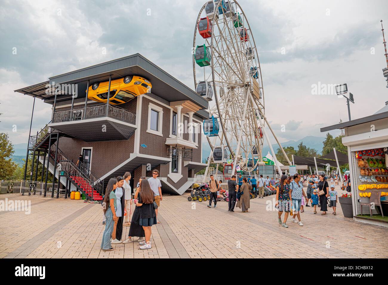22. Juni 2025, Almaty, Kasachstan: Menschen, die Freizeit im Vergnügungspark Kok Tobe genießen, mit umgedrehtem Haus und Riesenrad Stockfoto