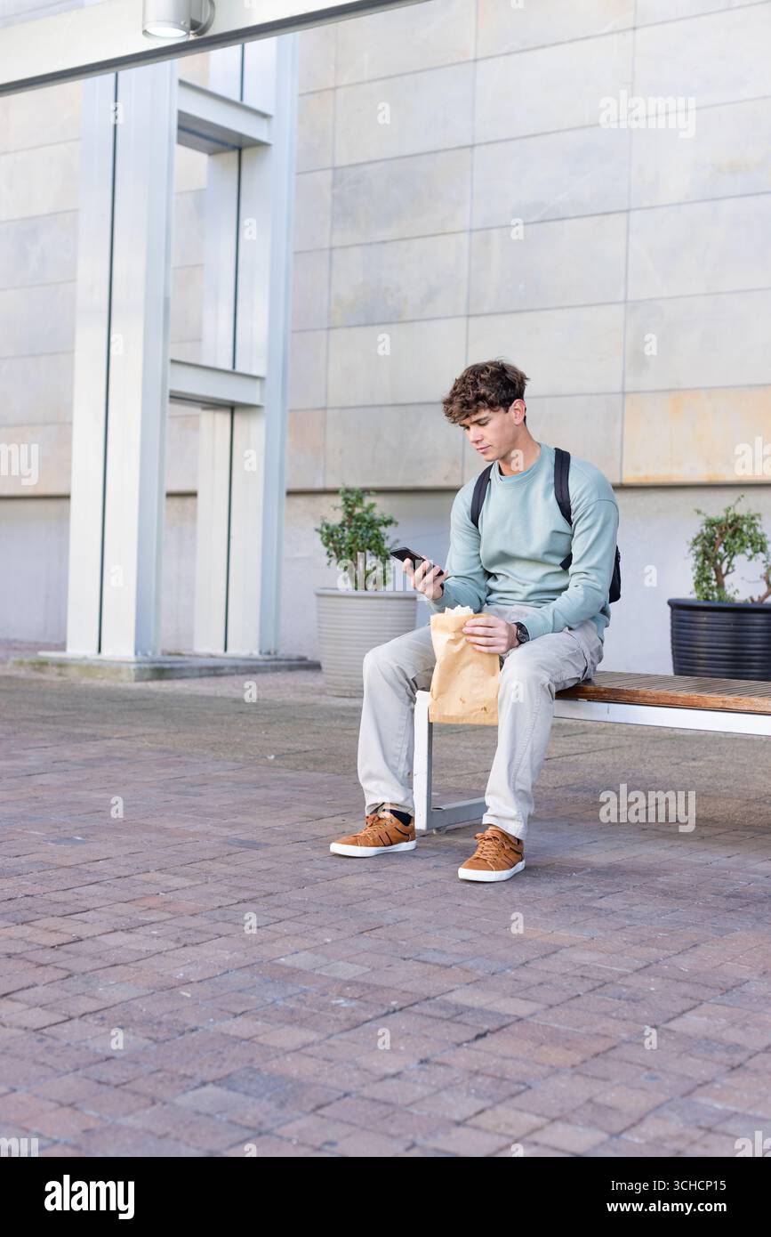 Männlicher Student sitzt auf einer Bank im städtischen plaza und hält Smartphone, Lunchtasche, Rucksack, Kopierraum. Lässig, Outdoor, Urban, modern, jugendlich, natürlich, c Stockfoto