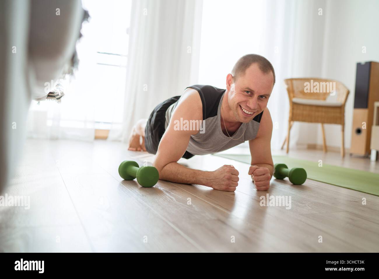 Home Workout im Wohnzimmer lächelnder junger muskulöser Mann, der Plank-Übung mit Kurzhanteln in der Nähe macht und einen gesunden Lebensstil fördert Fitness Motivat Stockfoto
