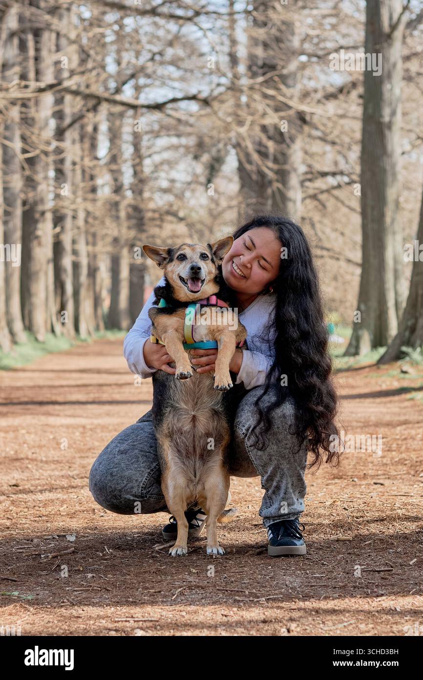 Die lächelnde lateinamerikanische Frau umarmt ihren Hund auf einem herbstlichen Waldweg und hält einen Moment der echten Verbindung in einer ruhigen natürlichen Umgebung fest. Stockfoto