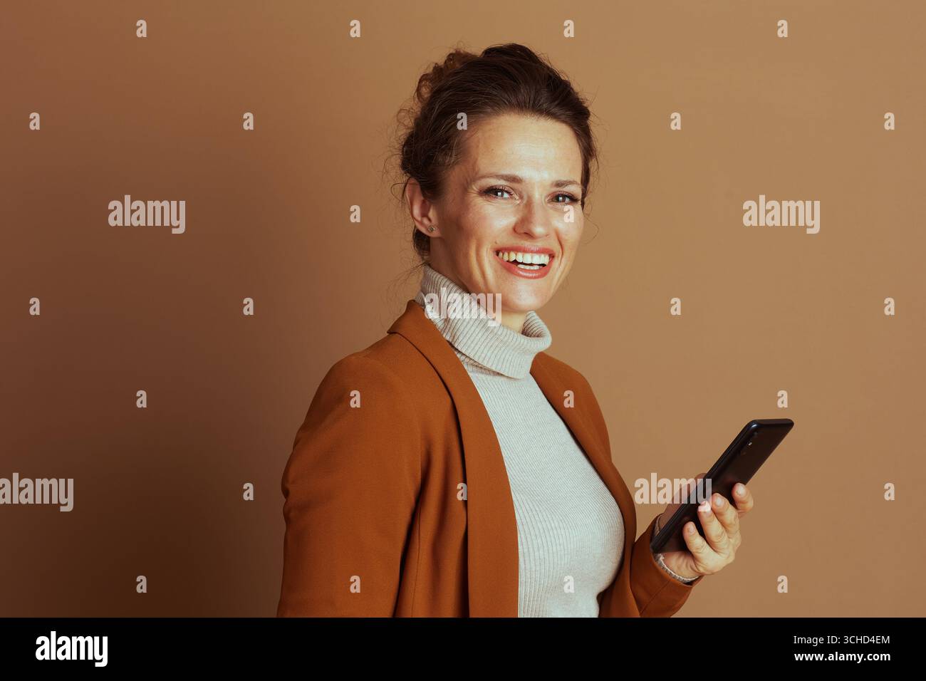 Eine selbstbewusste, lächelnde Geschäftsfrau in Blazer und Rollkragen hält das Smartphone und blickt direkt auf den Zuschauer. Dieses Porträt fängt den modernen Beruf ein Stockfoto
