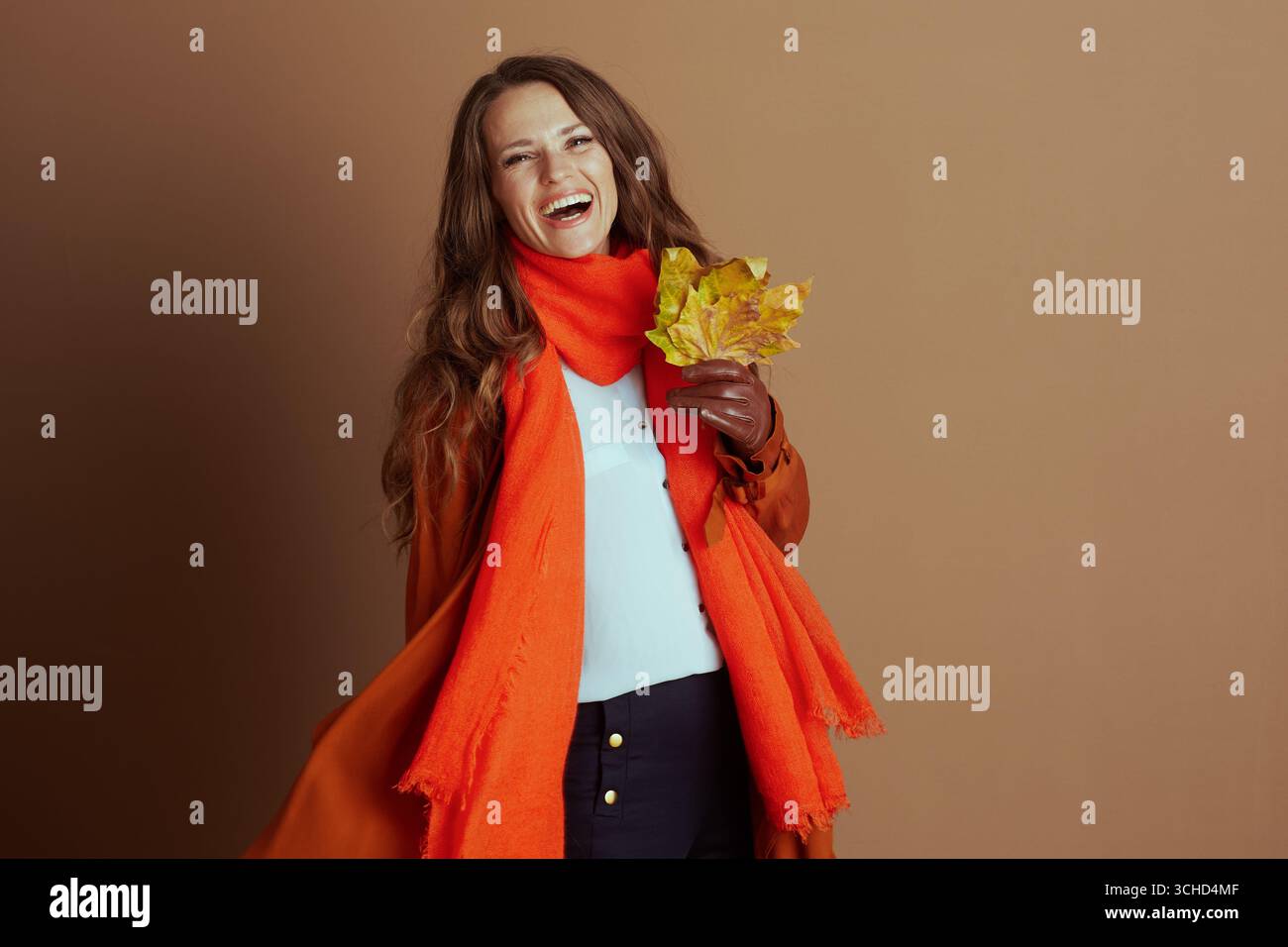 Eine fröhliche Frau mit gewelltem braunem Haar, gekleidet in einem leuchtend orangefarbenen Schal und Jacke, lacht herzhaft, während sie bunte Herbstblätter hält und die verkörpert Stockfoto