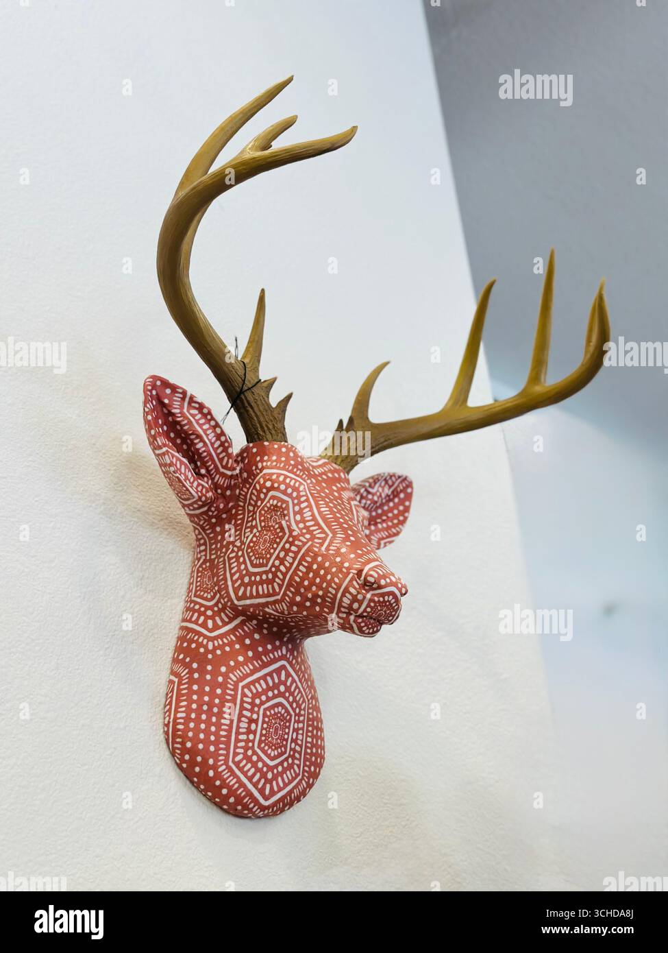 Handgefertigte Wandhalterung mit dekorativem Hirschkopf mit gestrichenem geometrischem Punktmuster, realistischem Geweih, an der weißen Wand drinnen. - Smartphone-aufgenommenes Stockfoto