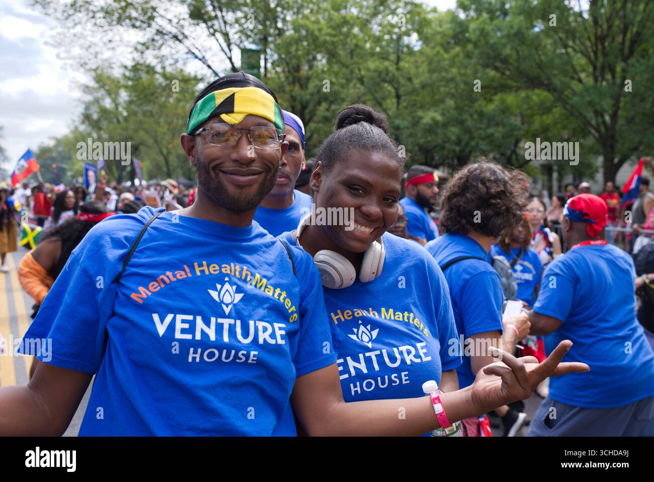 Zwei lächelnde Menschen in blauen T-Shirts bei einer Parade. Einer trägt ein Bandana mit jamaikanischer Flagge. Stockfoto