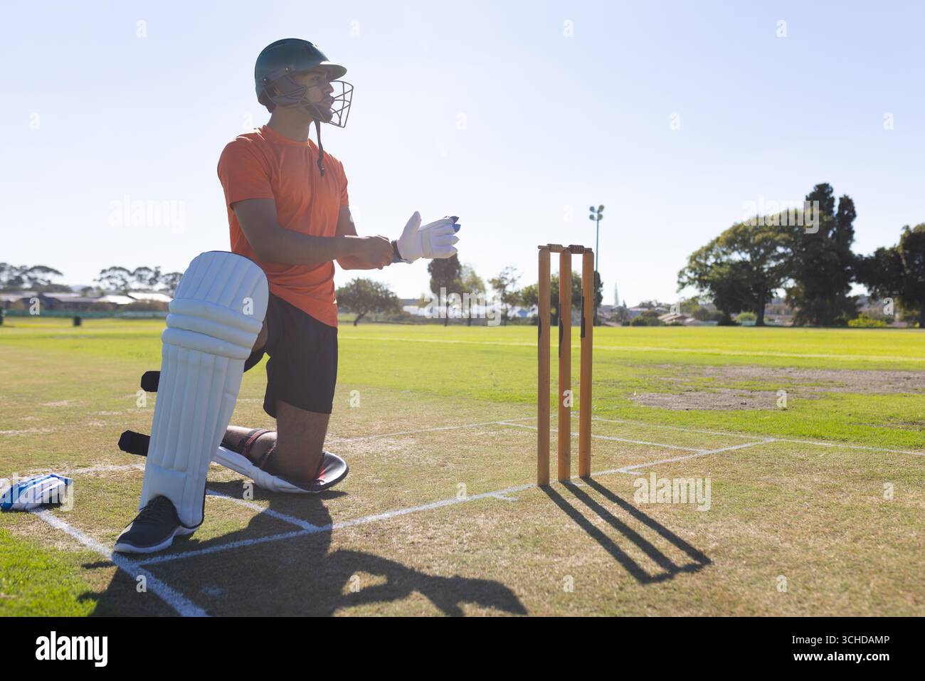 Cricketer in grünem Helm kniender Grasabstand verstellbarer Handschuhriemen durch Holzstümpfe Stockfoto