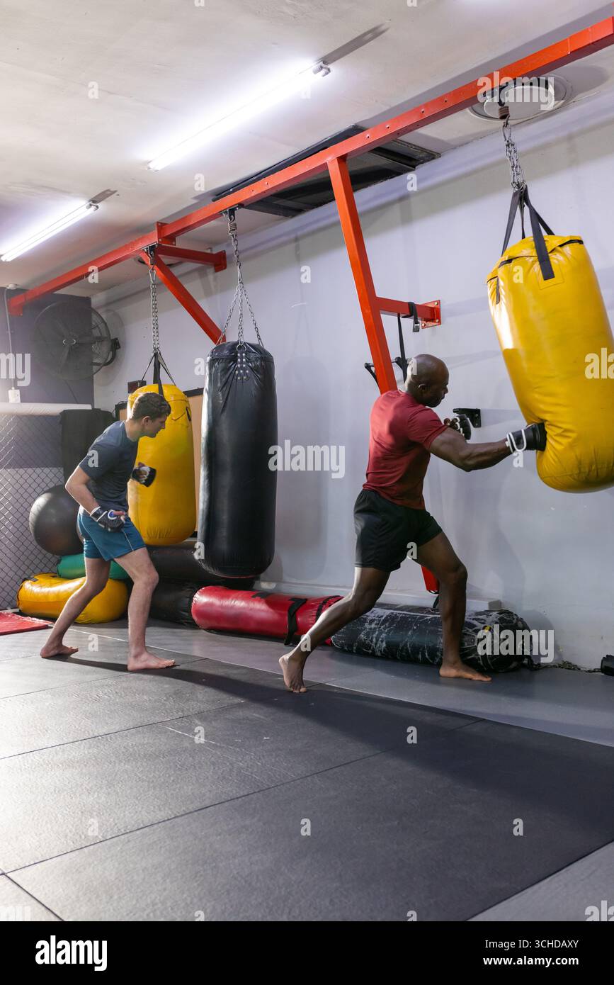 Verschiedene männliche Boxpartner in Shorts, die gelbe, schwarze schwere Taschen im Boxstudio schlagen Stockfoto