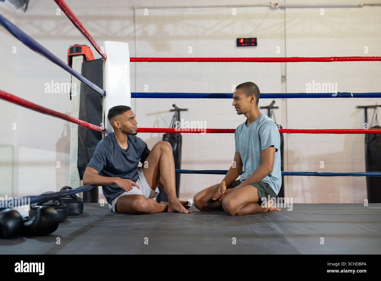 Zwei männliche Trainingspartner sitzen auf einer Ringmatte im Boxring neben Kettlebells, Timer Stockfoto