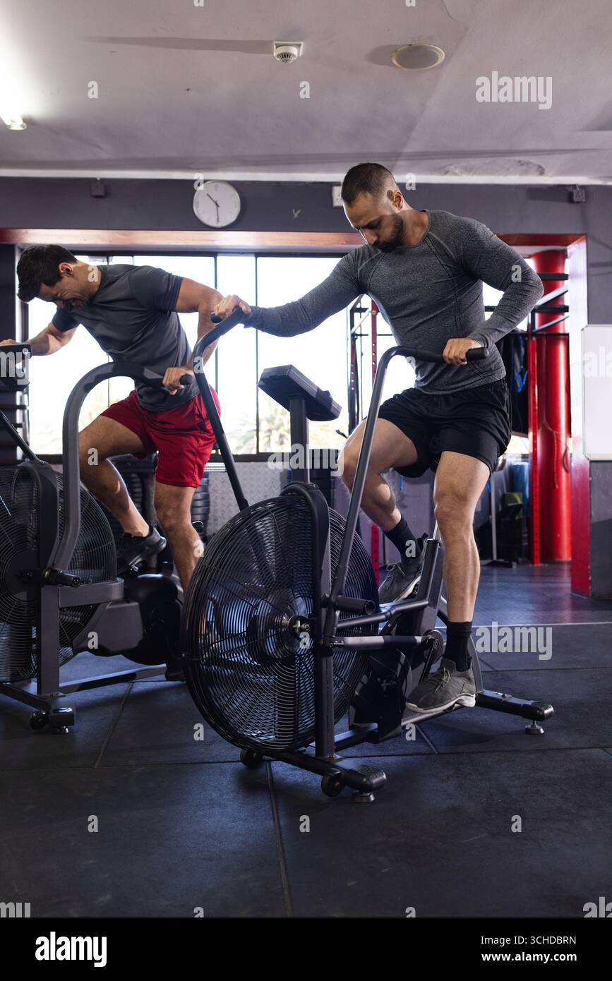 Verschiedene männliche Trainingspartner, die stationäre Luftfahrräder in der Turnhalle in Sportbekleidung in die Pedale treten Stockfoto