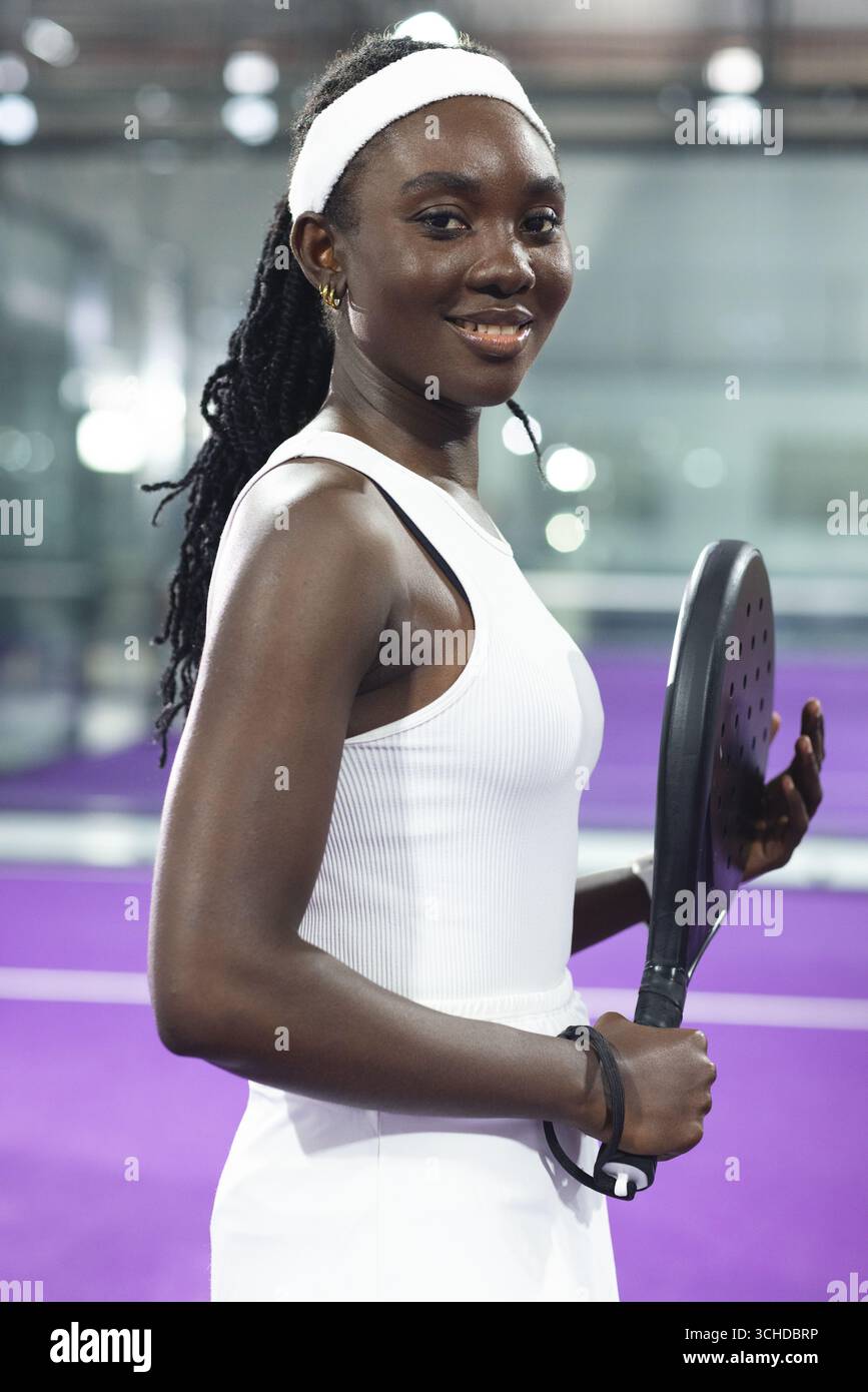afroamerikanerin, die drinnen auf dem purpurpurfarbenen Padel-Hof mit Schläger und Stirnband steht Stockfoto