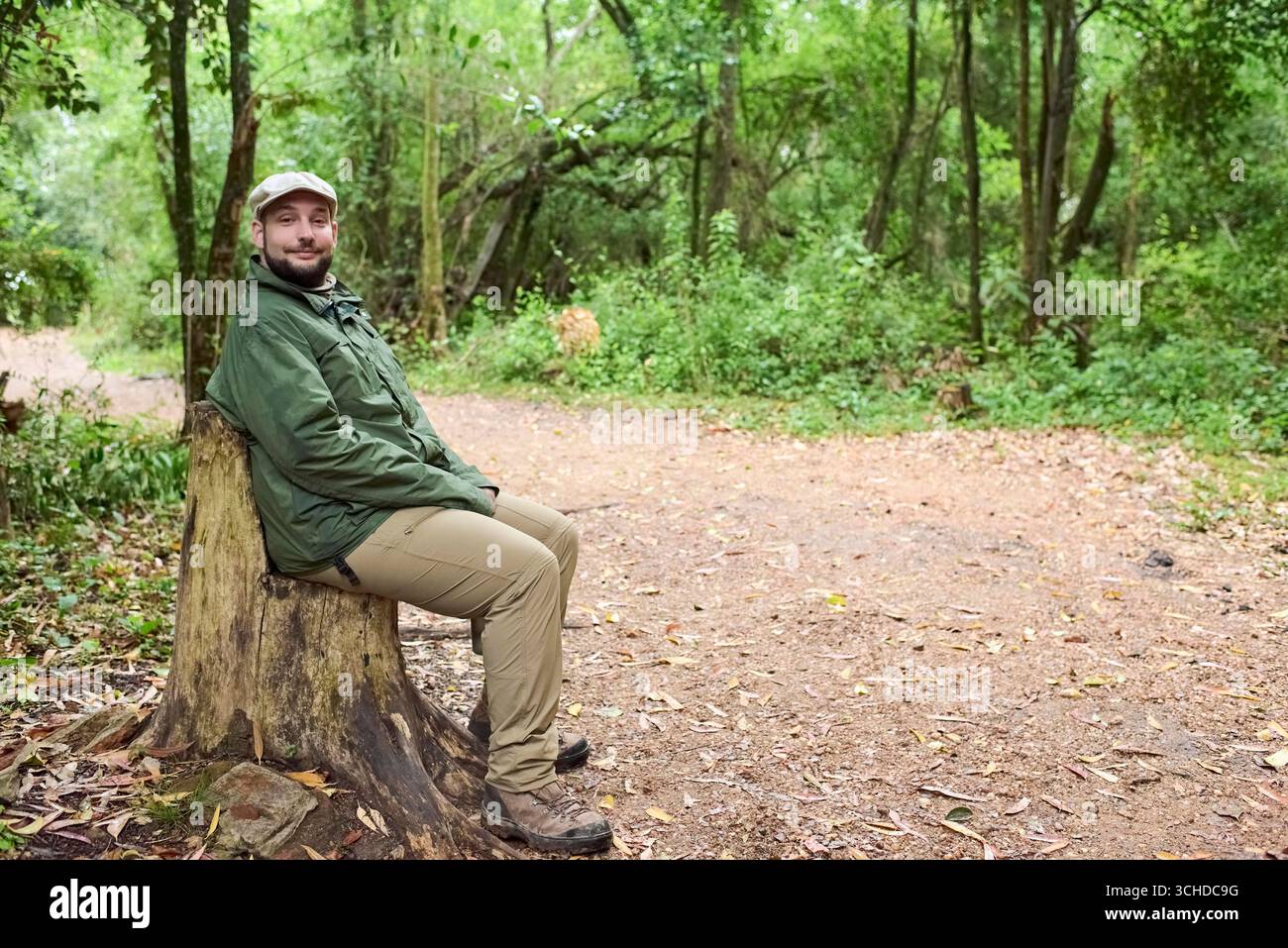 Junger hispanischer Mann, der auf einem Baumstumpf sitzt, als Sitz neben einem Wanderweg in einer natürlichen Umgebung, El Palmar Nationalpark, Entre Rios, Argentinien. Stockfoto