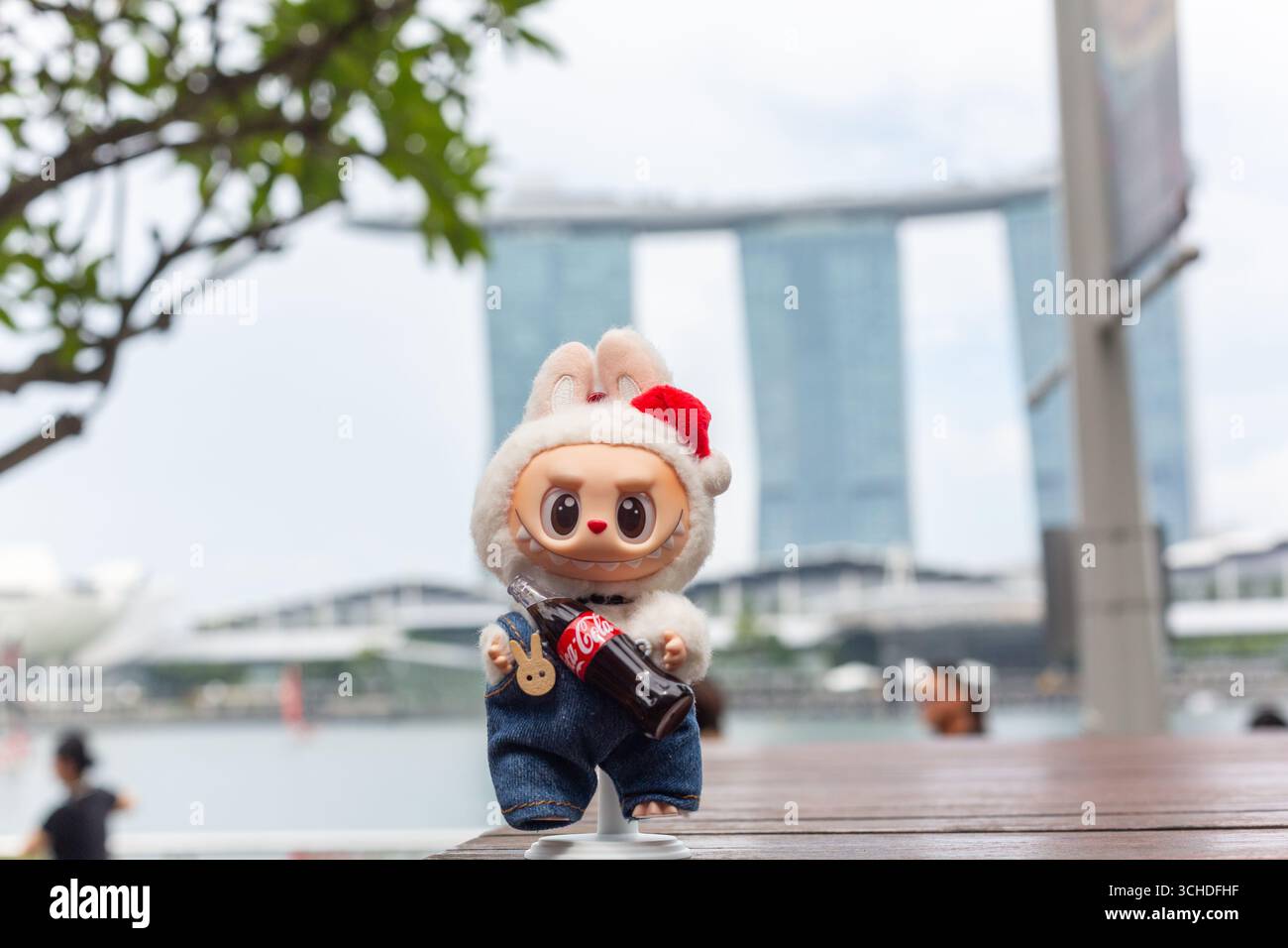 Weiße Labubu-Figur mit einer Cola-Flasche, die sich vor dem Marina Bay Sands Hotel posiert, ein Wahrzeichen in Singapur. Stockfoto