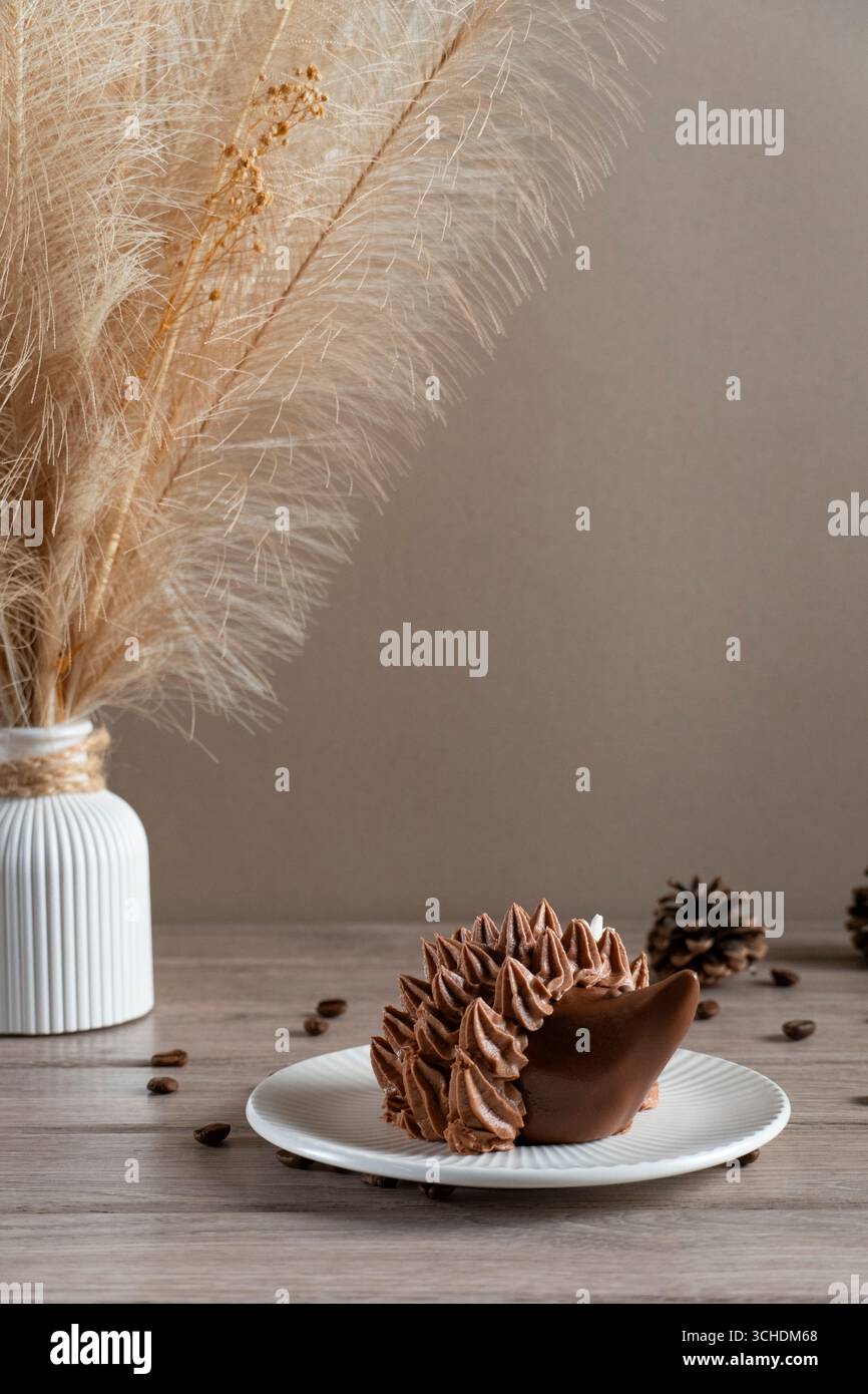Süßes Schokoladen-Igel-Dessert auf einem weißen Teller mit rustikalem Pampas-Gras in Vase und Tannenzapfen auf hölzernem Hintergrund. Gemütliche Herbst- oder Winterdesserts sc Stockfoto