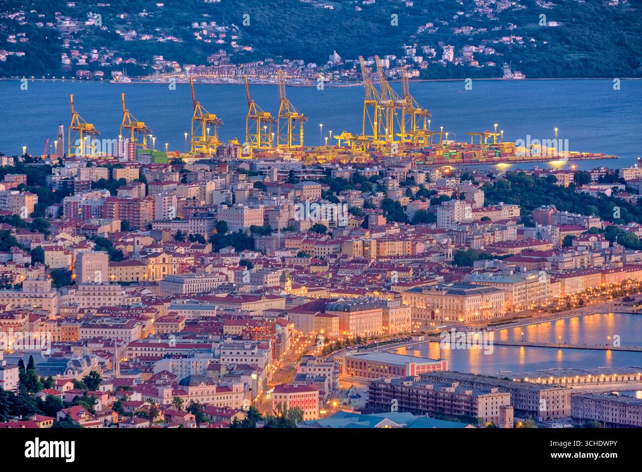 Aus der Vogelperspektive auf die Häuser, den Yachthafen und die Kräne des Überseehafens von Triest, beleuchtet bei Nacht. Stockfoto