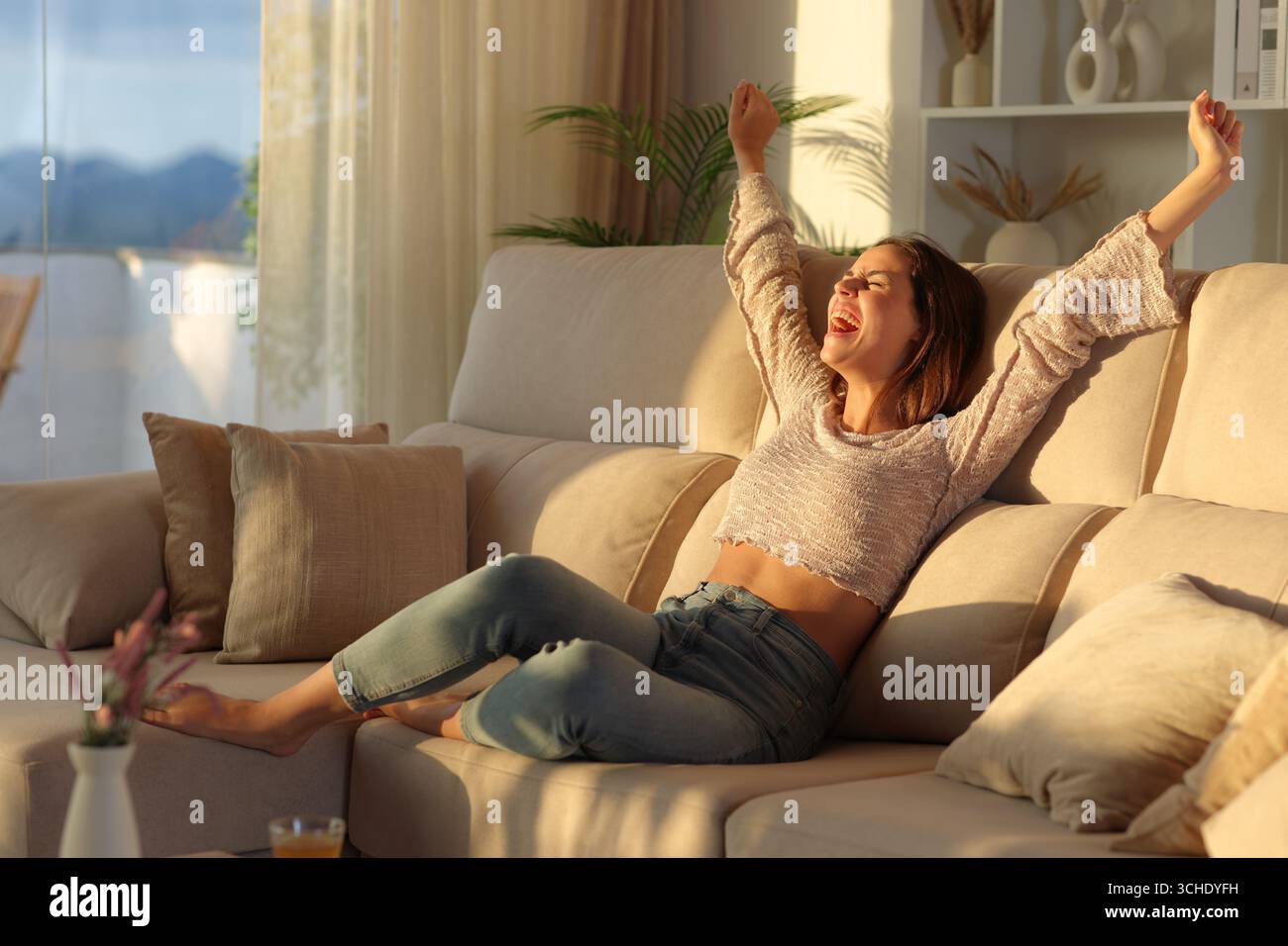 Aufgeregte Frau, die bei Sonnenuntergang die Arme hebt und zu Hause auf einer Couch sitzt und feiert Stockfoto