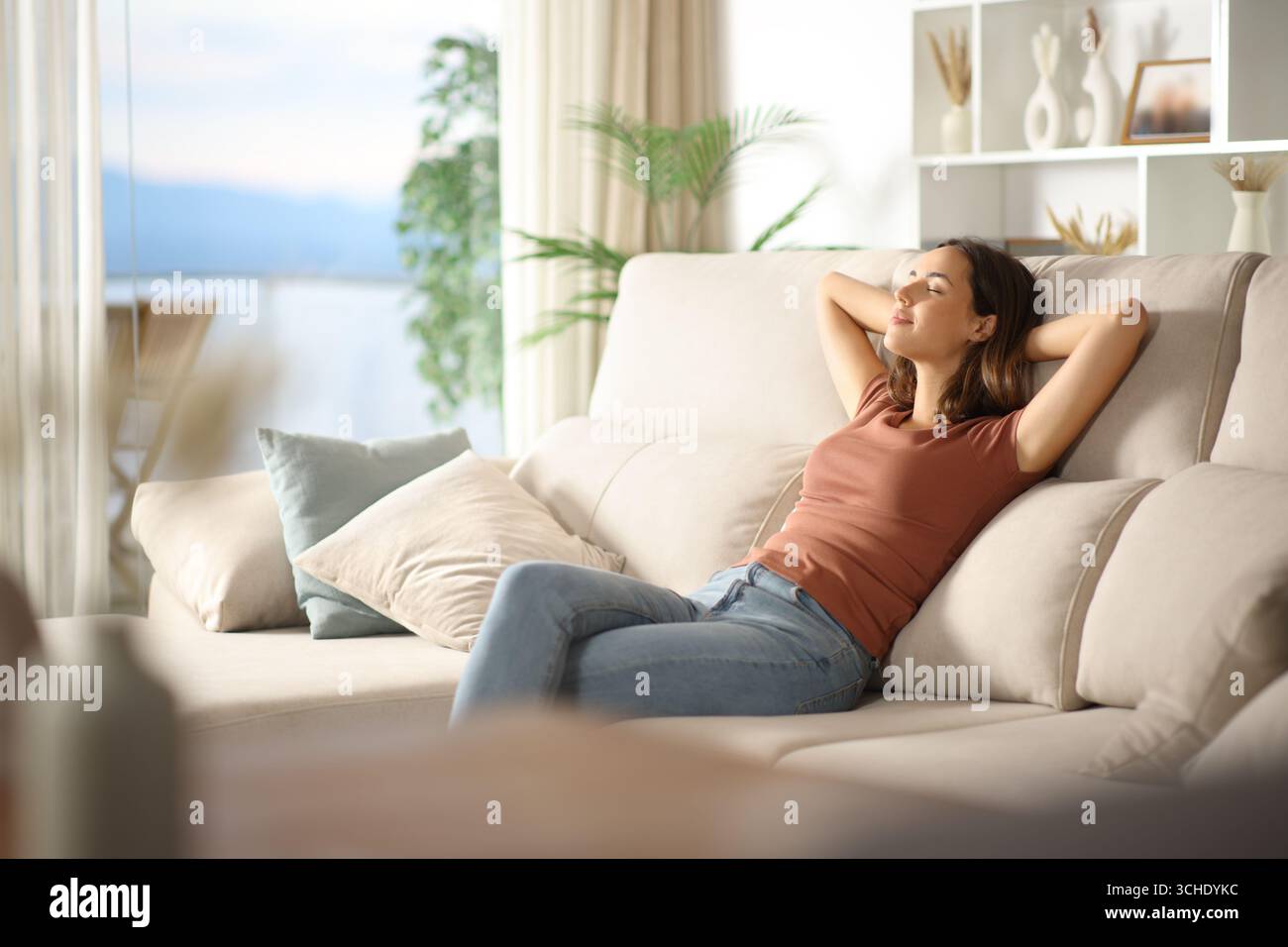 Frau, die auf einem Sofa sitzt und sich zu Hause neben einem Fenster entspannt Stockfoto