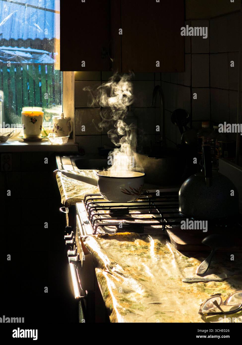 Sonnenlicht strömt durch ein Fenster auf einen Topf mit dampfenden warmen Speisen, die auf einem Herd in einer gemütlichen Küche zubereitet werden, und schafft eine warme und einladende Atmosphäre Stockfoto