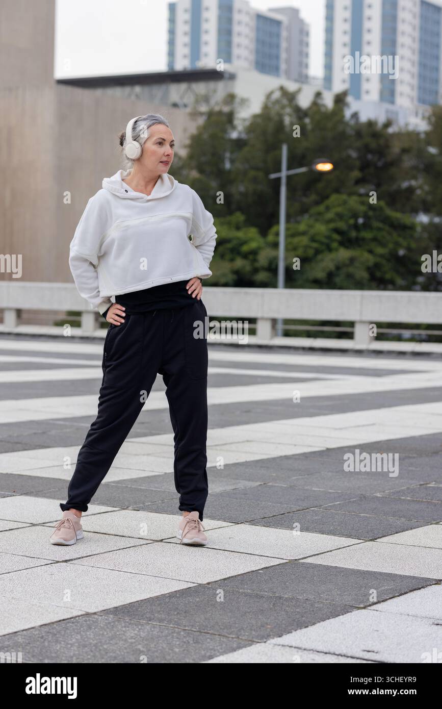 Reife Frau, die auf dem fußgängerplatz neben Betongeländer trainiert, mit Kopfhörern Stockfoto