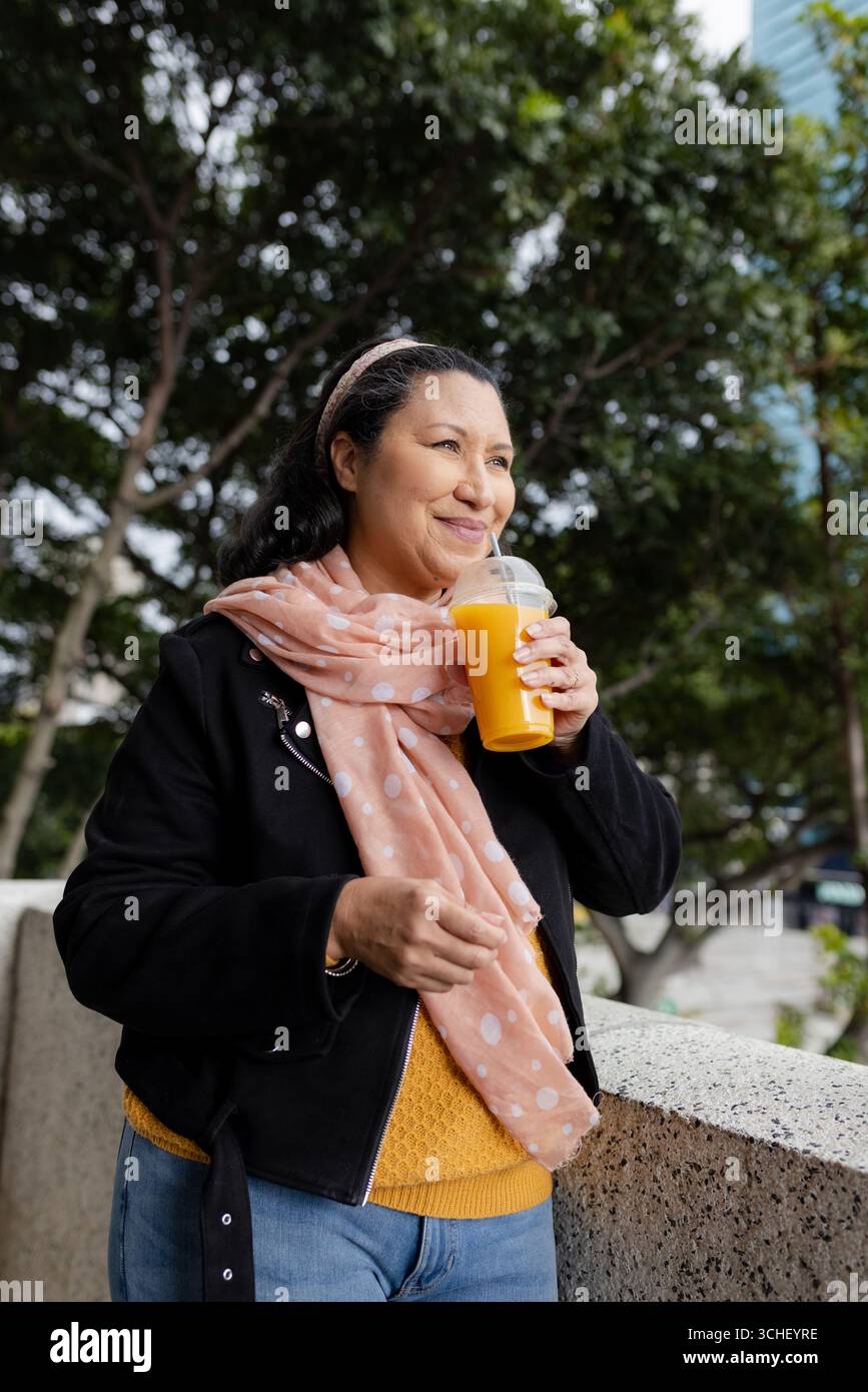Hispanische Frau lehnt sich an Betongeländer auf dem städtischen platz, mit Orangensaftbecher mit Strohhalm Stockfoto