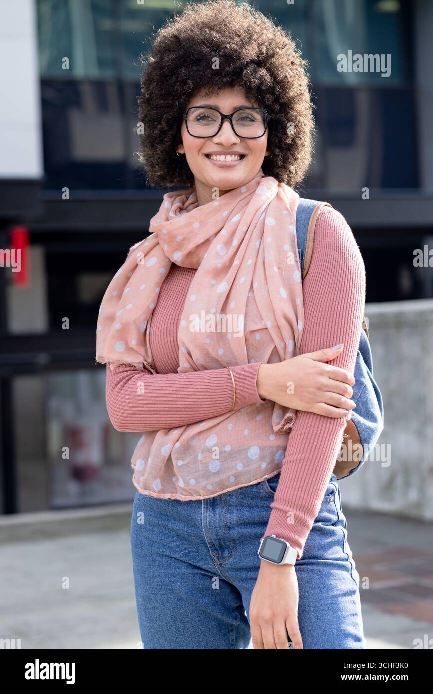 afroamerikanerin, die auf dem plaza vor dem Glasgebäude steht, in Schal, Brille, Rucksack Stockfoto