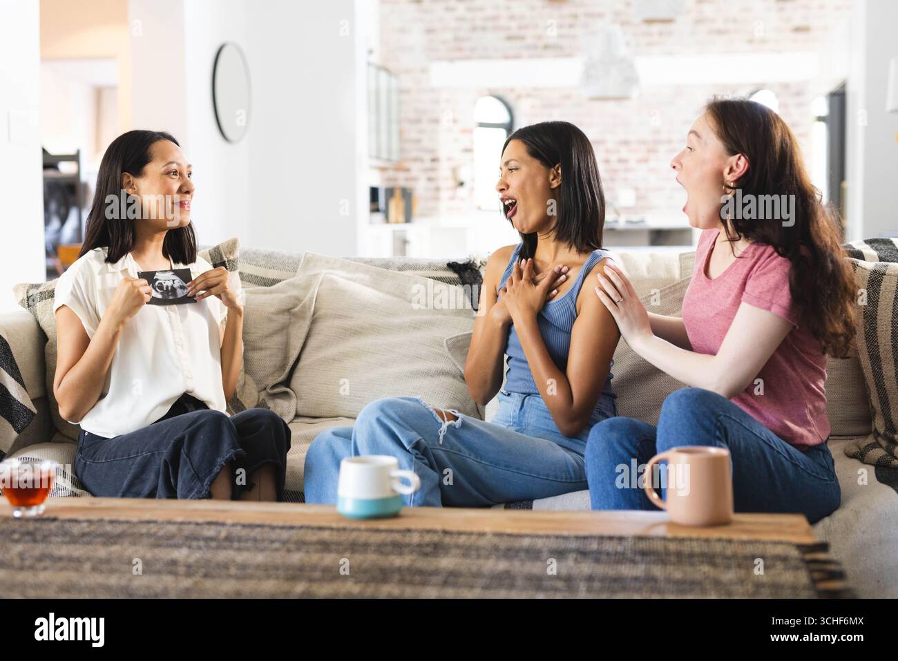 Verschiedene Freundinnen reagieren fröhlich auf spannende Nachrichten, während sie zu Hause auf einem gemütlichen Sofa sitzen Stockfoto