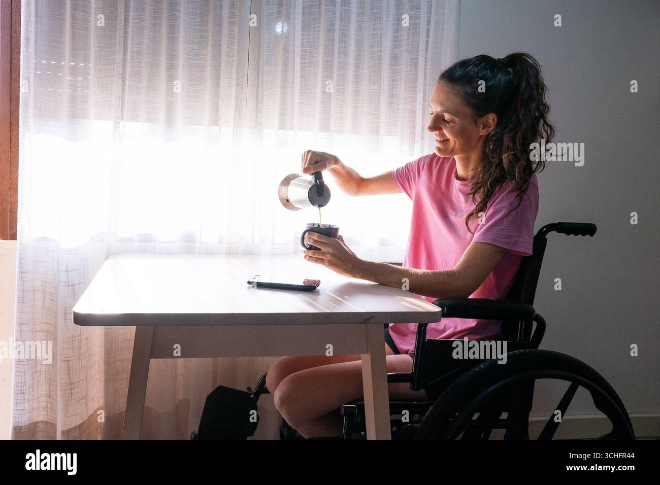 Glückliche Frau mit Behinderung genießt eine Tasse Kaffee zu Hause, gießt Kaffee aus einer Mokakanne, fördert unabhängiges Leben und Inklusivität Stockfoto