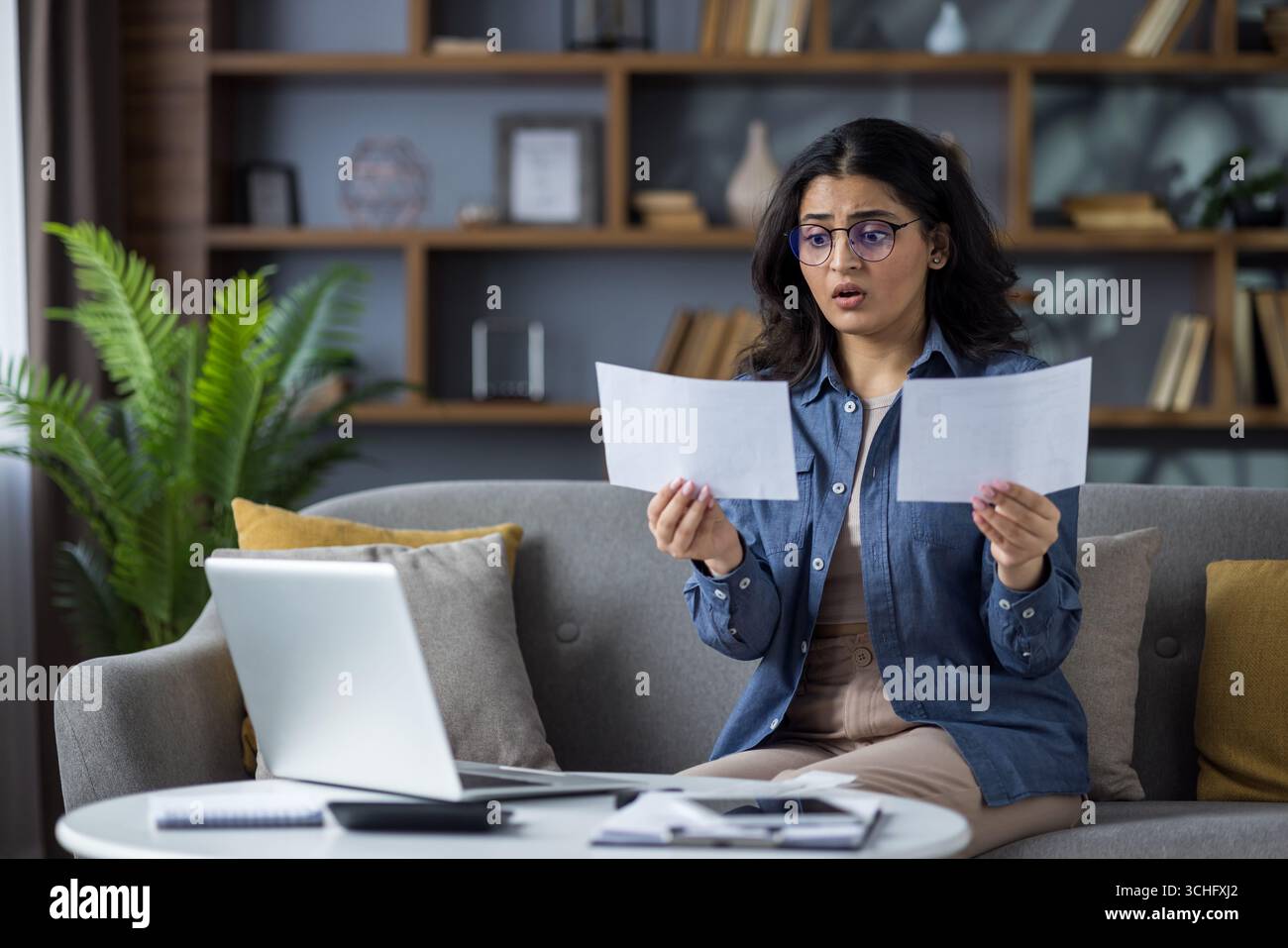 Besorgte junge Indianerin sitzt zu Hause auf der Couch und sieht schockiert auf die Dokumente aus, die sie hält. Stockfoto