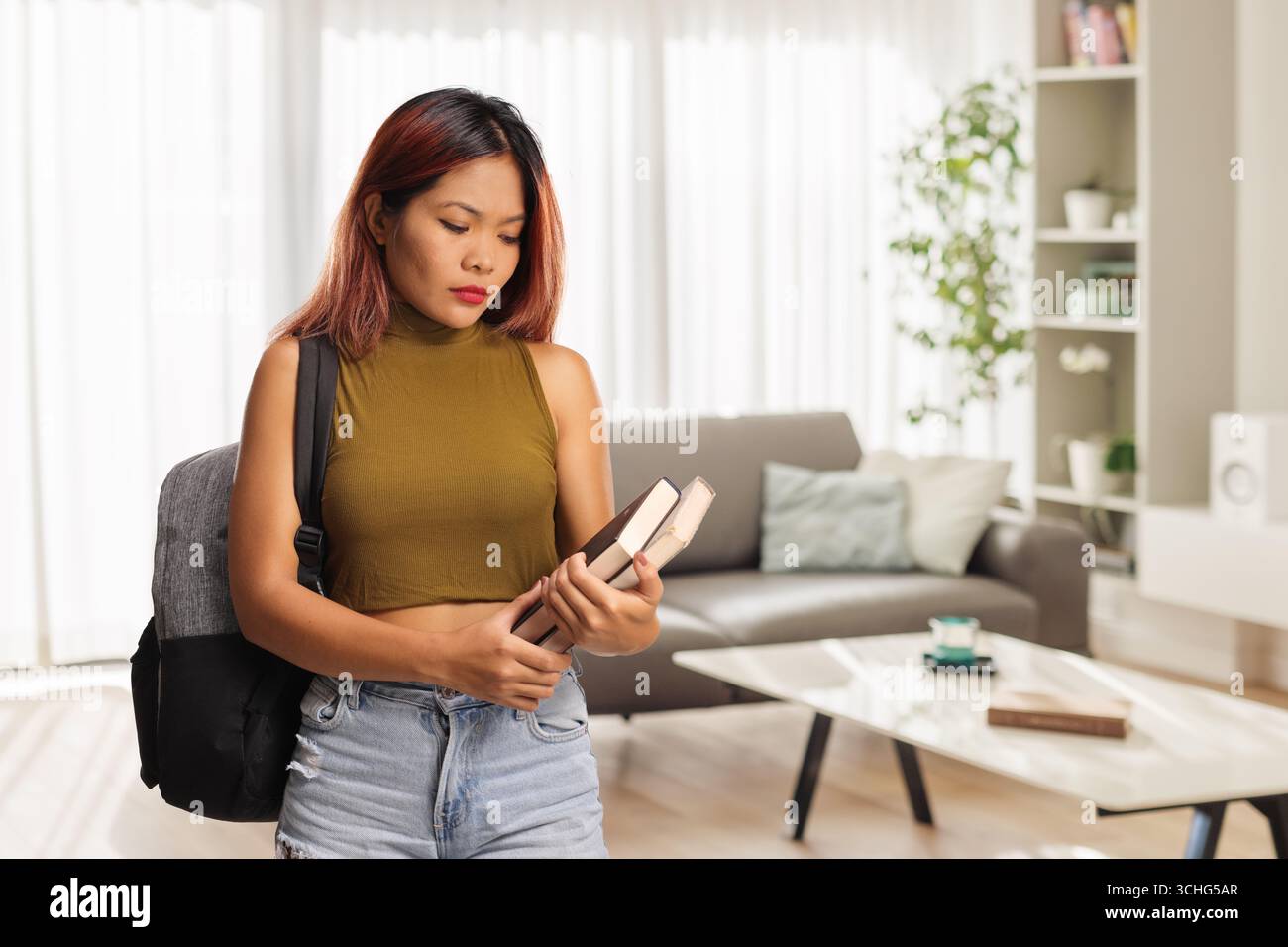 Nachdenkliche junge Frau, die Bücher hält und im Wohnzimmer steht Stockfoto