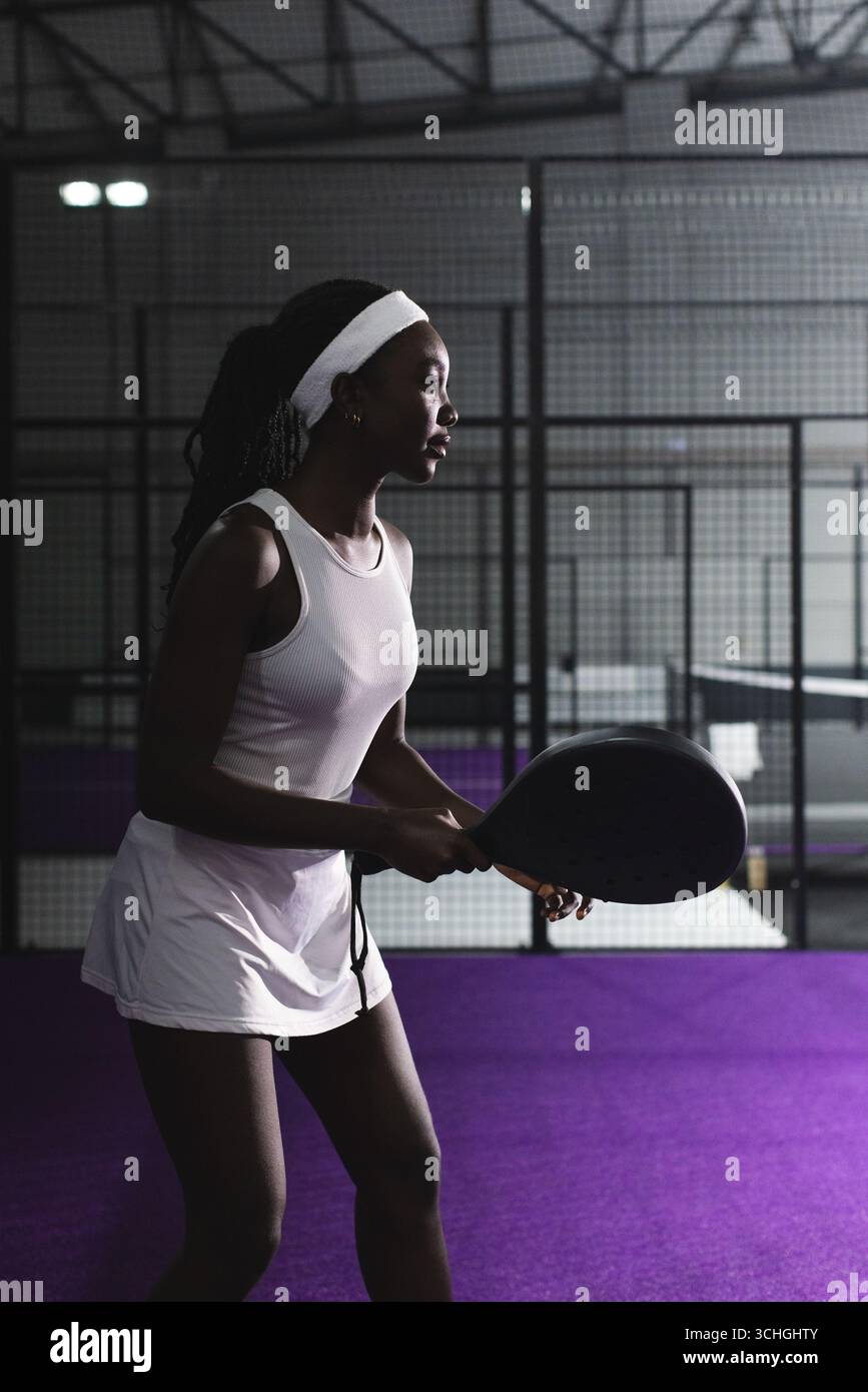 Weibliche Athletin mit Padel-Schläger auf dem violetten Spielfeld im Käfig unter hellen Deckenleuchten Stockfoto