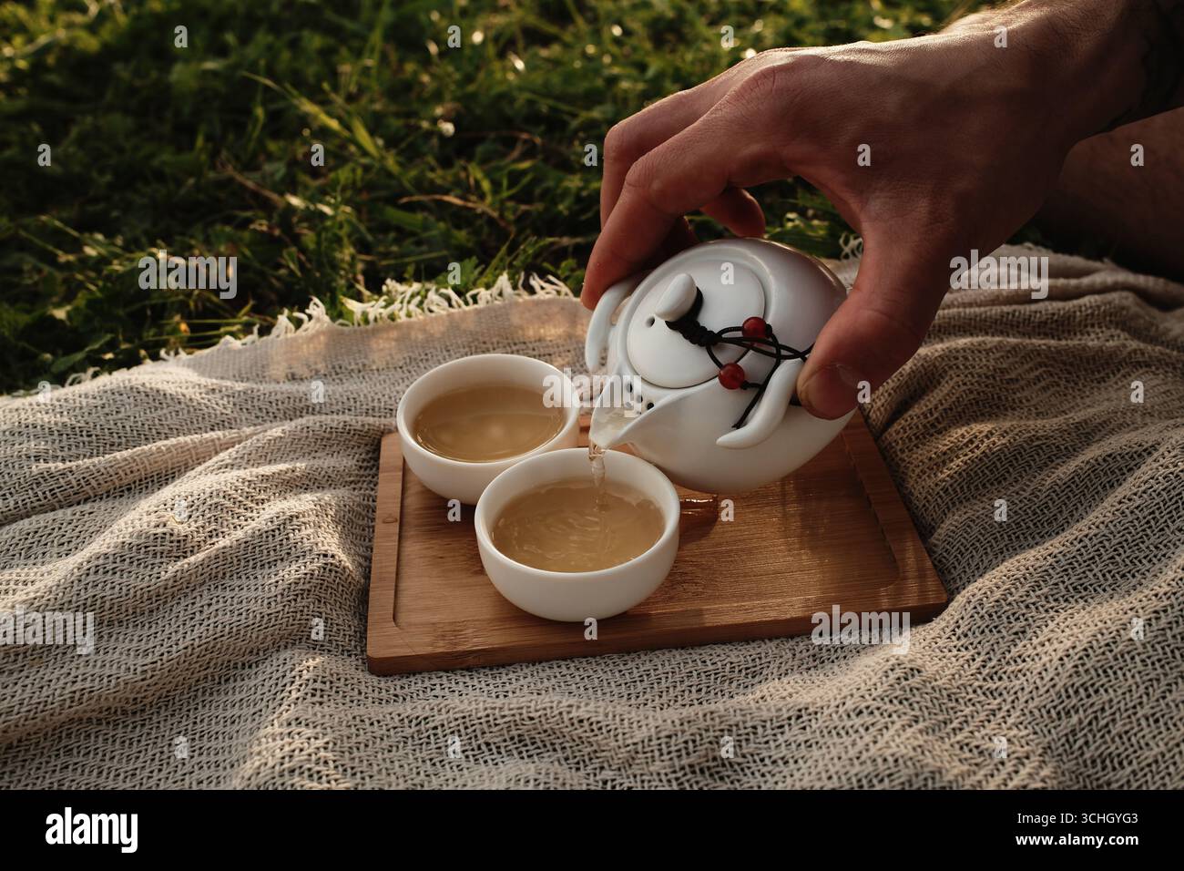 Geben Sie heissen Tee von Hand aus einer weißen Teekanne in eine kleine Porzellantasse draußen auf einem Holztablett. Picknick im chinesischen Stil für zwei Personen im Freien. Stockfoto