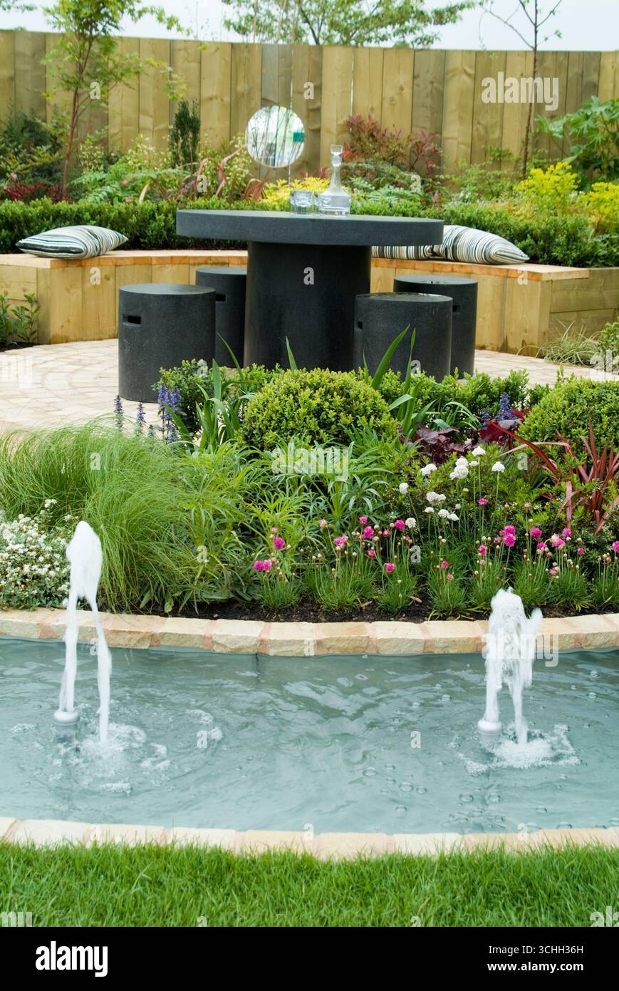 Ein ruhiger Außenbereich zeigt einen modernen Tisch mit Sitzgelegenheiten, umgeben von farbenfrohen Pflanzen und einem ruhigen Wasserspiel, perfekt zum Entspannen. Stockfoto