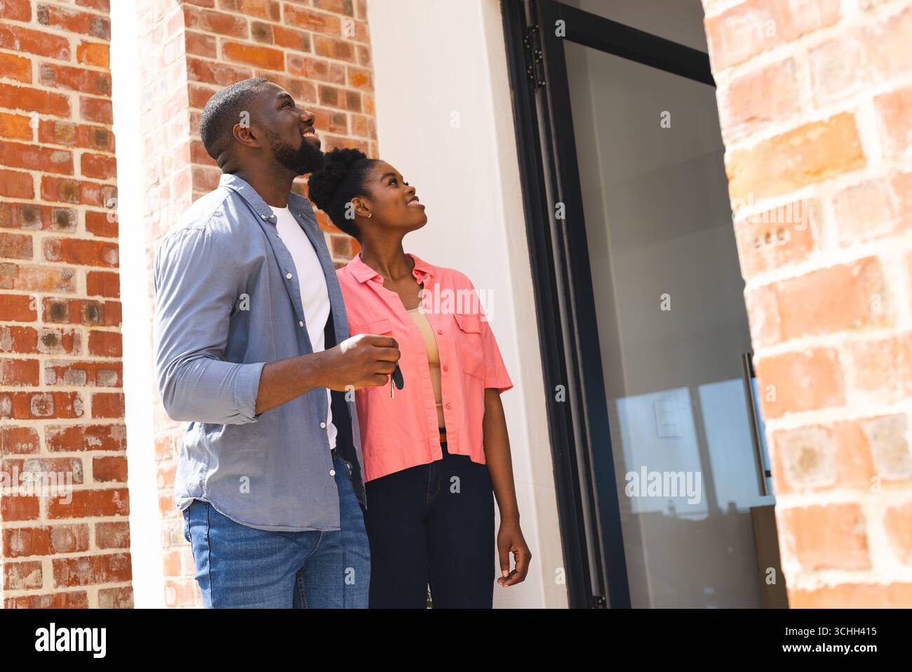 Glückliches Paar, das Schlüssel hält, vor dem neuen Zuhause steht, aufgeregt und hoffnungsvoll aussieht Stockfoto