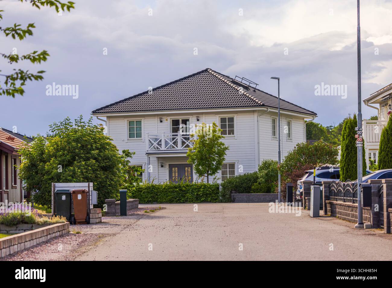 Modernes zweistöckiges weißes Haus mit dunklem Dach, umgeben von Bäumen und Büschen in einer ruhigen Vorstadtstraße. Schweden. Stockfoto