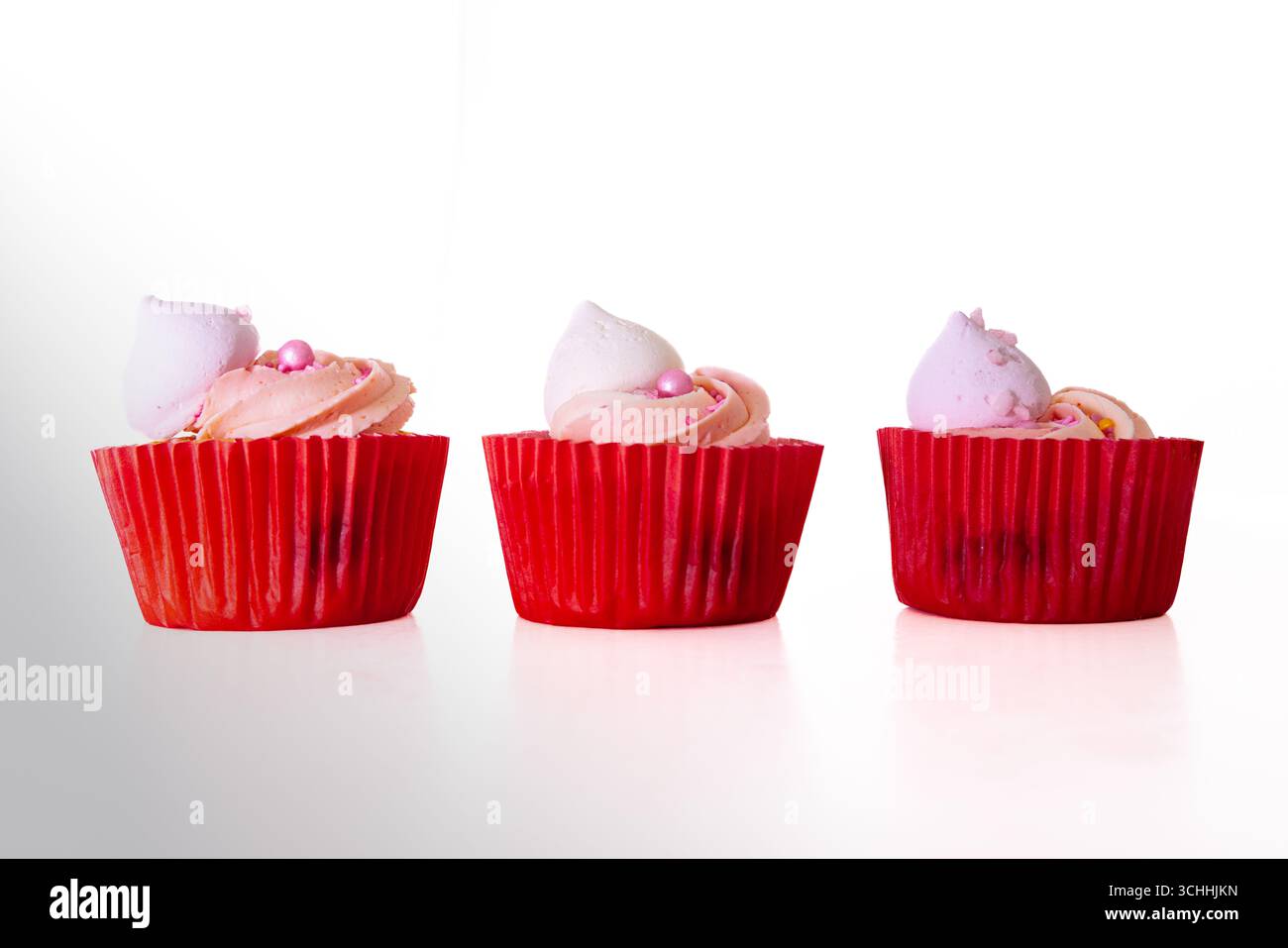 Drei rosafarbene Cupcakes mit verdrehtem Frosting, Meringue-Topper und Zuckerperlen in roten Verpackungen, nebeneinander auf weißem Hintergrund angeordnet. Stockfoto