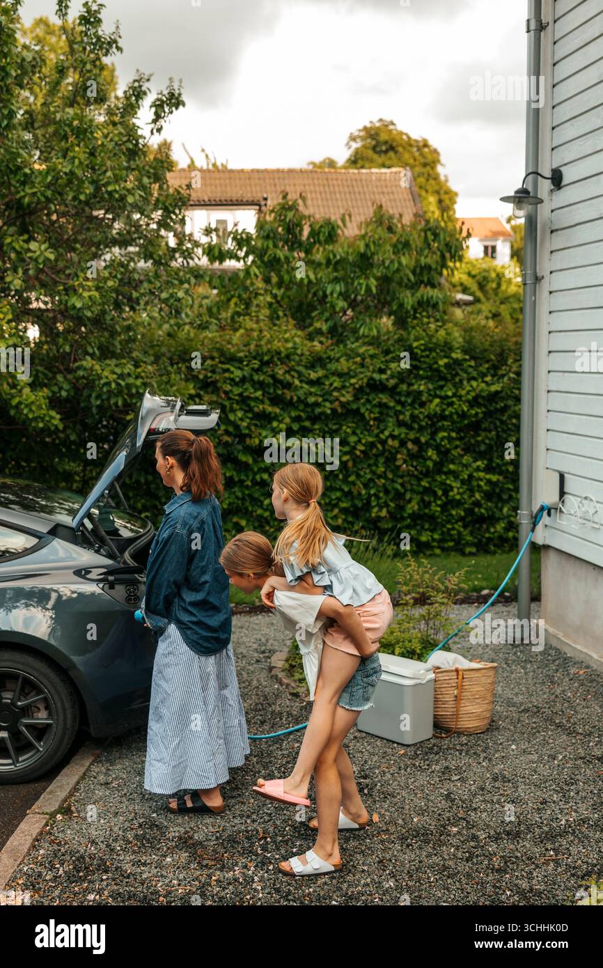 Mädchen, die Schwester, die in der Nähe der Mutter steht und ein Elektroauto im Garten auflädt Stockfoto