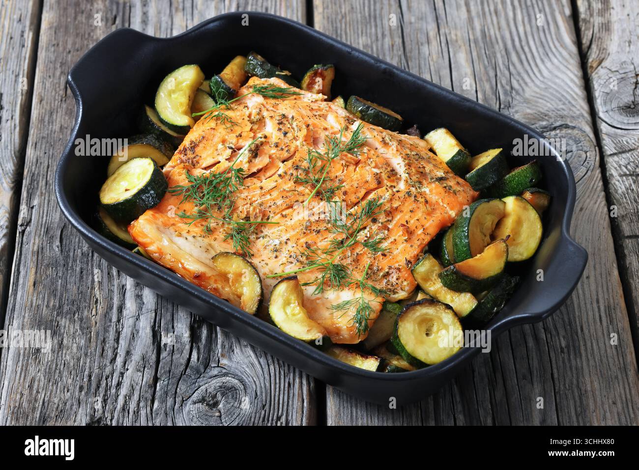 Gebacken im Ofen Lachsfilet mit Zucchini und Dill in schwarzer Auflaufform auf rustikalem Holztisch, horizontale Ansicht, Nahaufnahme, nicht KI generiert Stockfoto