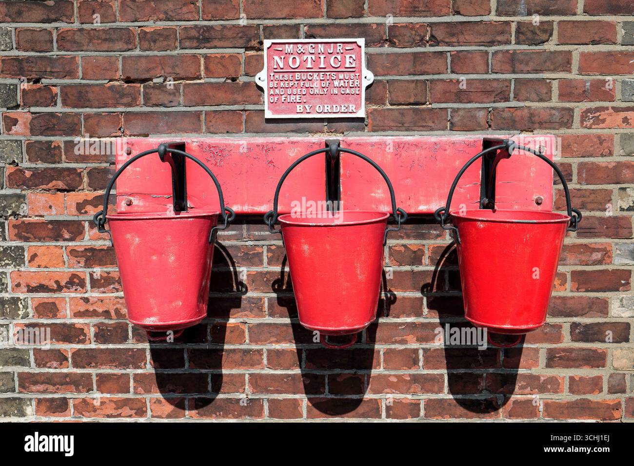 Drei rote Feuereimer hängen an einem Wandregal am Holt Railway Station an der North Norfolk Railway (Poppy Line) Stockfoto