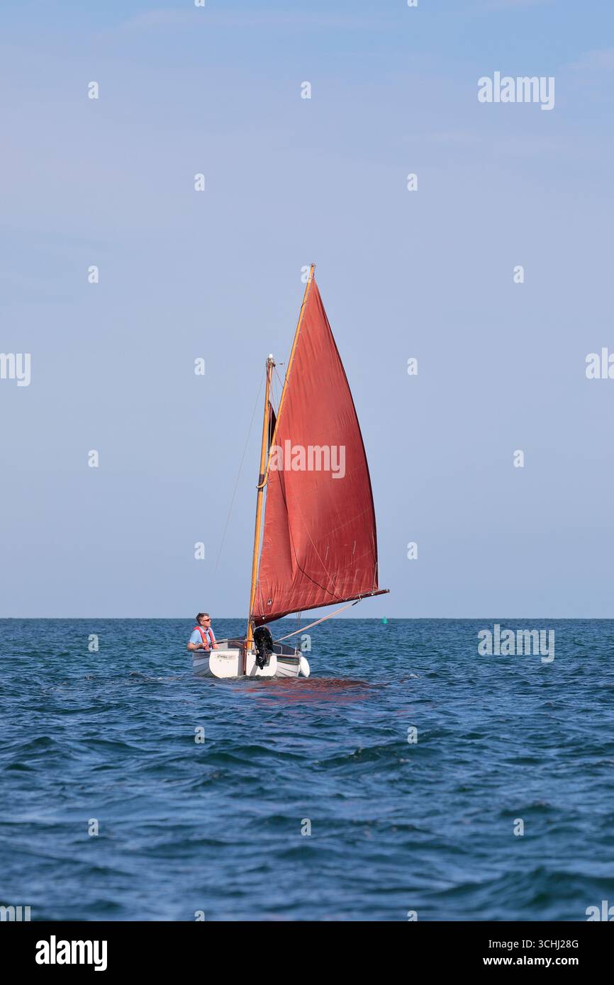Ein kleines grün-weißes Segelboot namens „Catcher“ mit roten Segeln und einem Außenbordmotor, der auf dem offenen Meer segelt Stockfoto