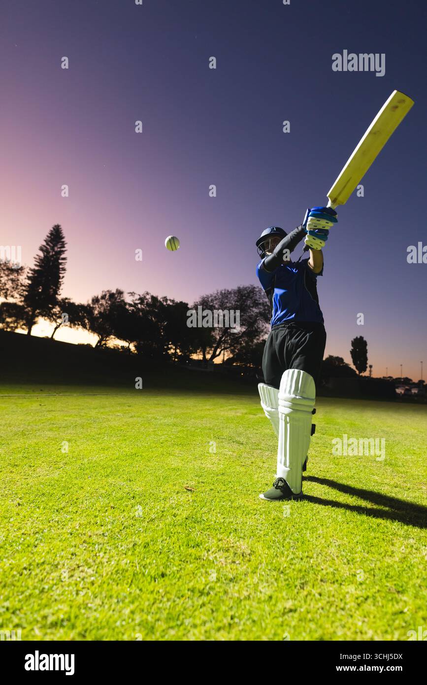 Indischer Cricketspieler schwingt Schläger bei grünem, weißem Ball auf dem Grasfeld in der Dämmerung Stockfoto